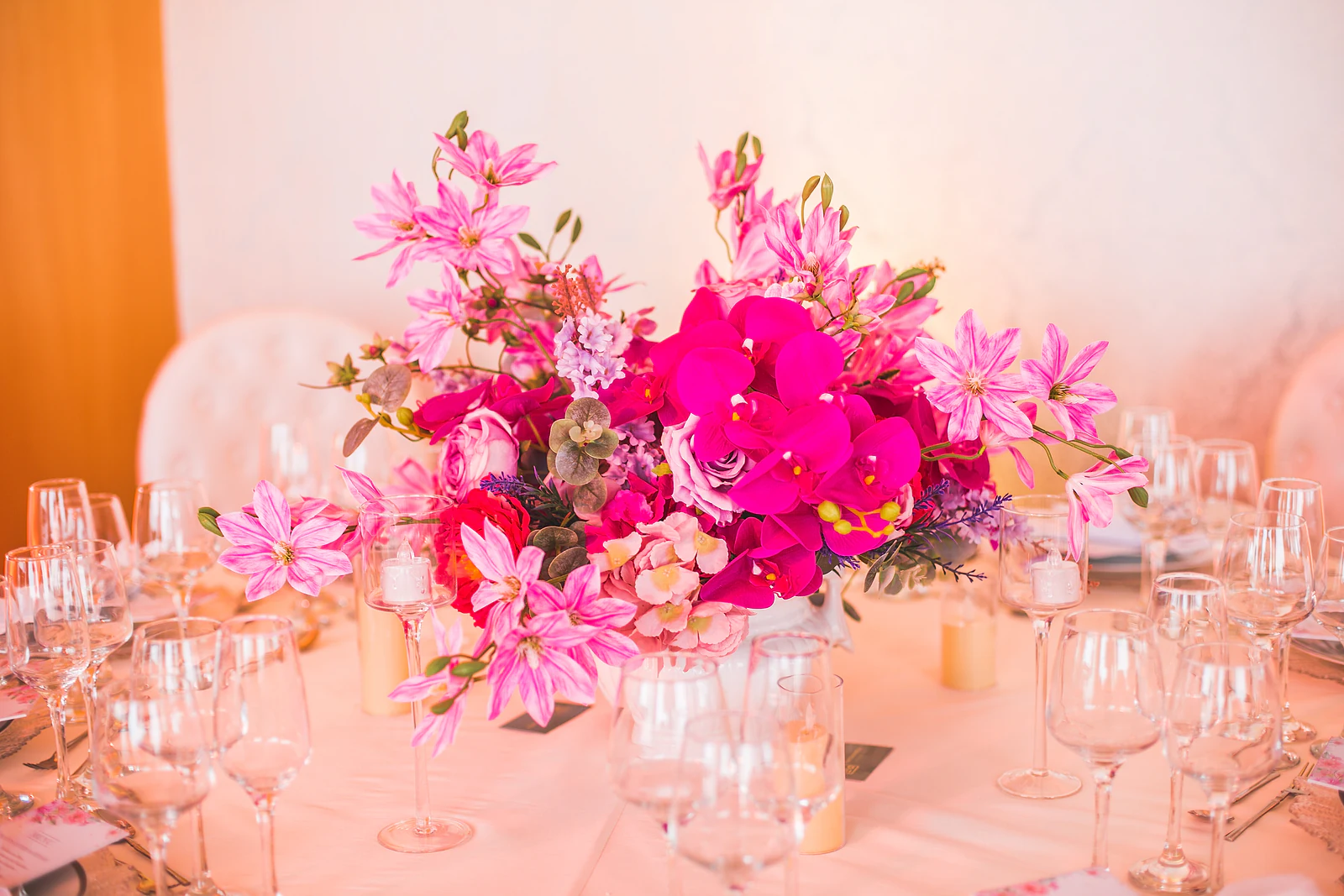 Centro de mesa baixo de orquídeas fúcsia, rosas e flores lilás entre copos de cristal e velas