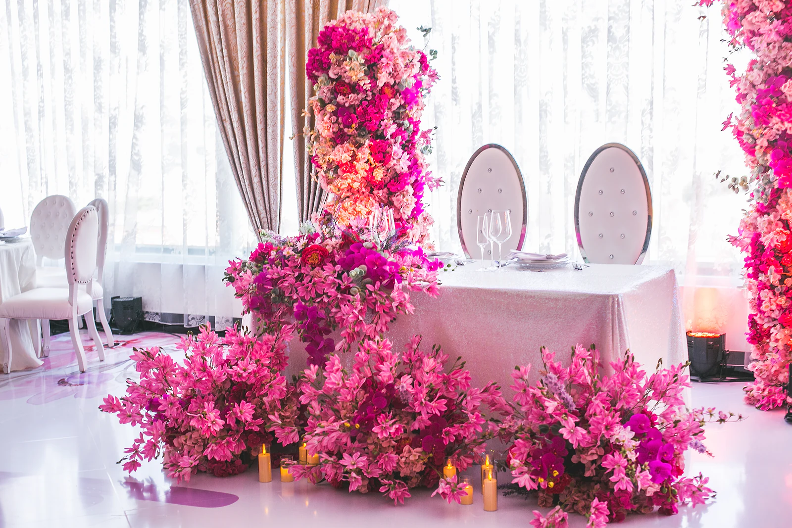 Mesa dos noivos de toalha cintilante, com duas cadeiras de espaldar oval, uma cascata de flores cor-de-rosa e velas no chão
