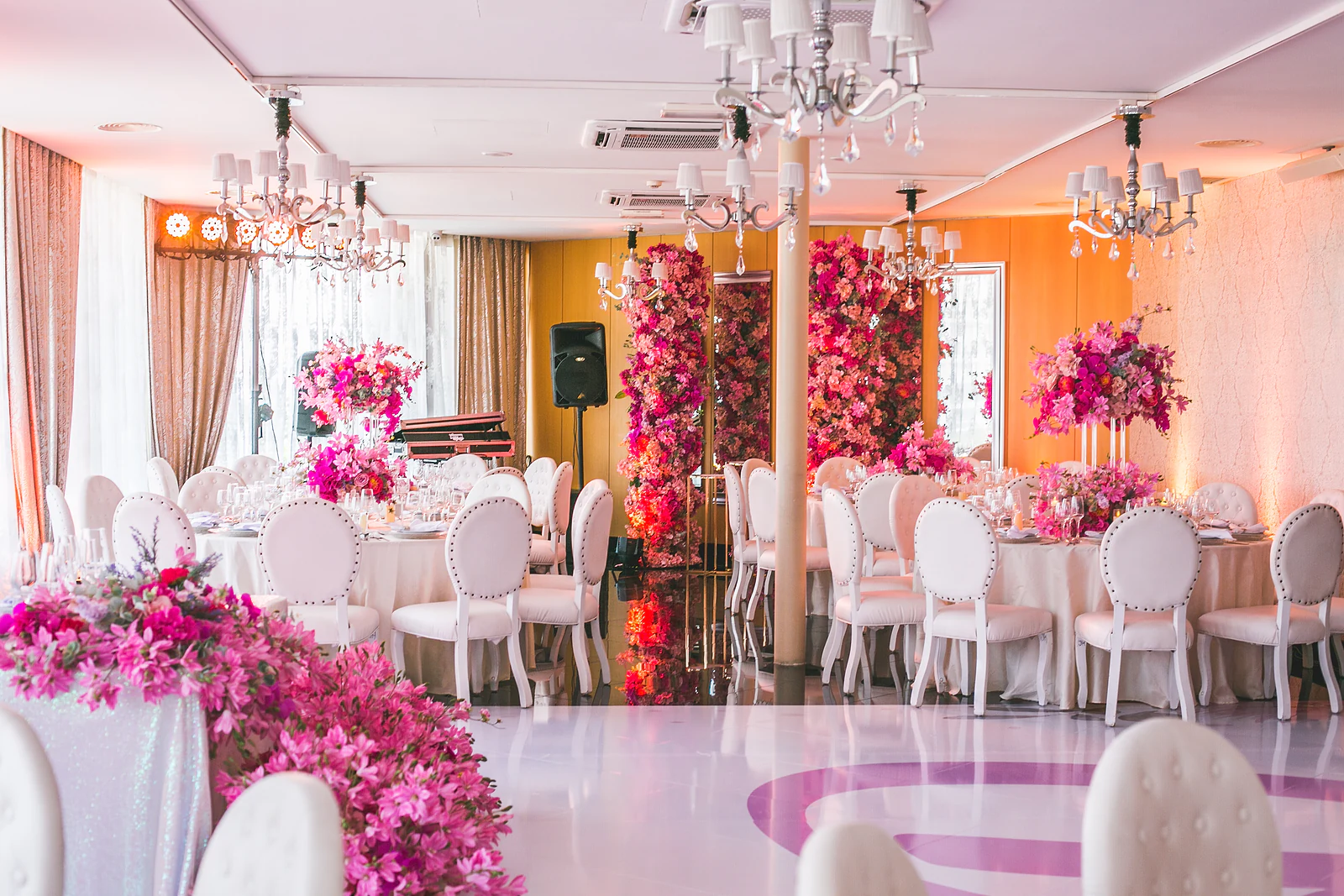 Sala vista da pista, com uma cascata de flores cor-de-rosa em primeiro plano, mesas redondas de cadeiras brancas, colunas de flores ao fundo e lustres