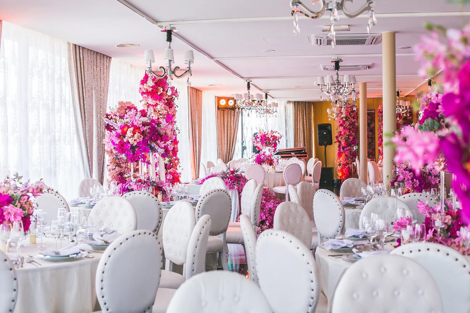 Sala com mesas redondas, cadeiras brancas almofadadas, colunas de flores fúcsia e lustres, sob cortinados claros