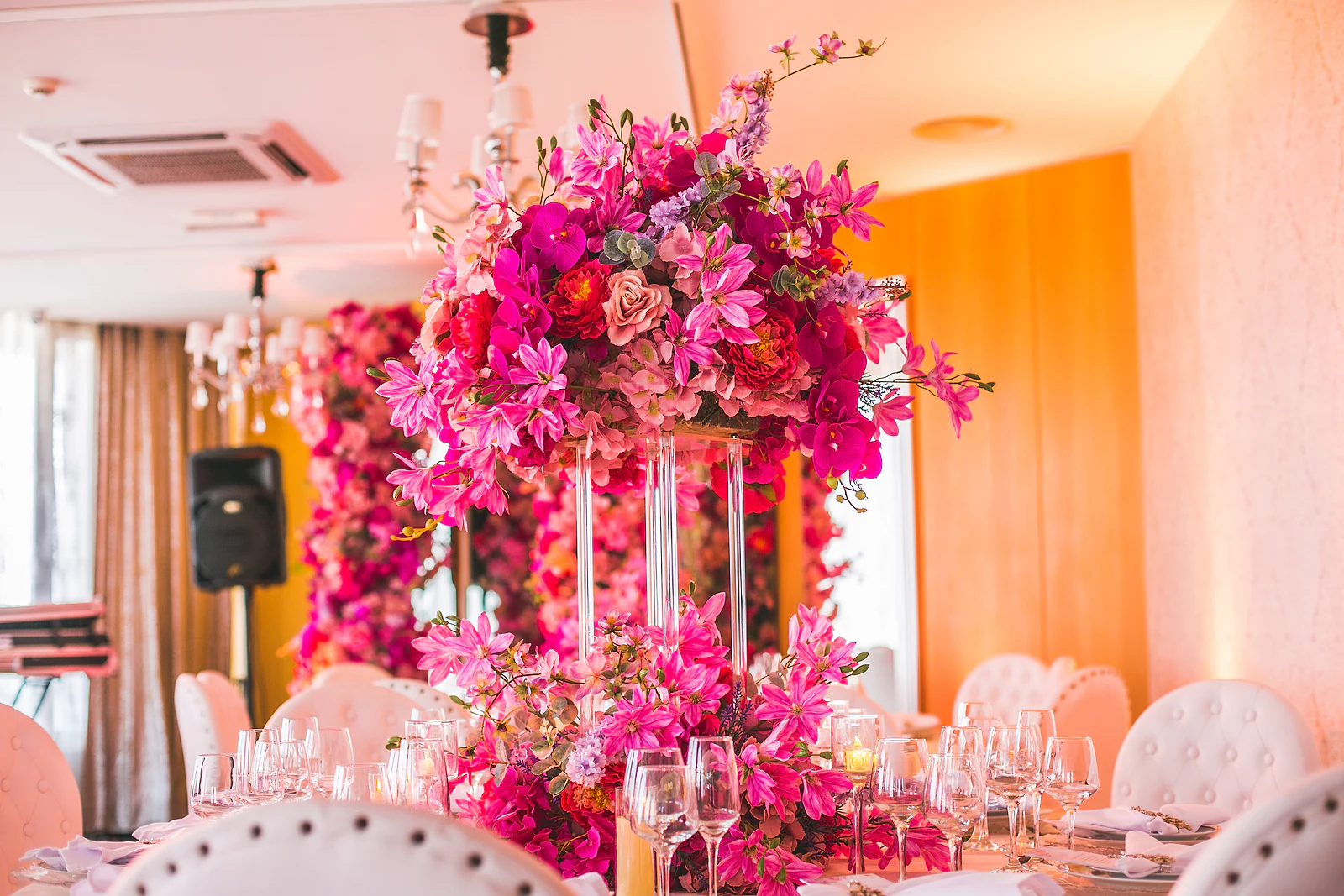 Arranjo alto de orquídeas e flores cor-de-rosa sobre colunas de vidro, com lustres e uma parede de flores ao fundo