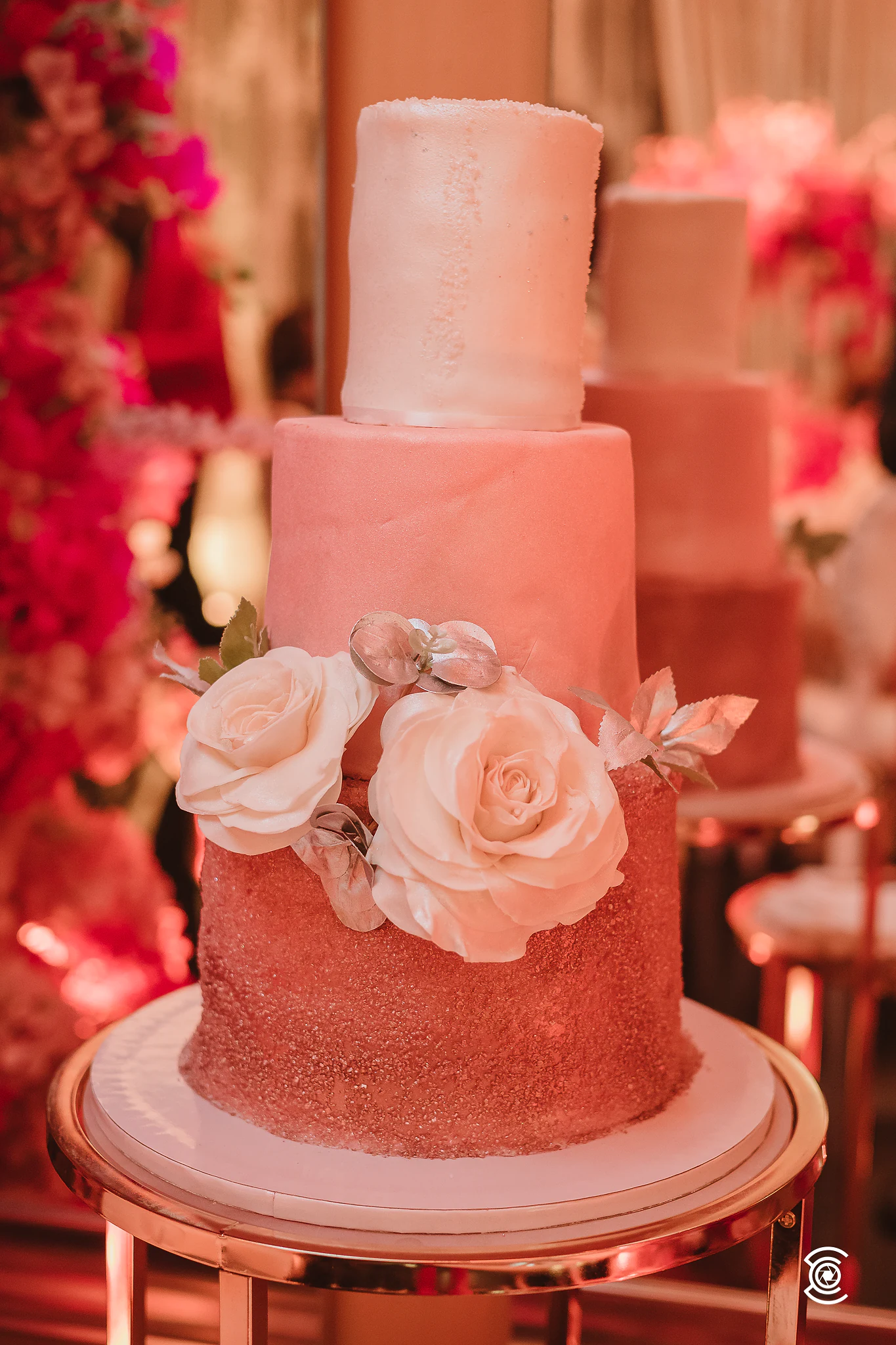 Bolo de três andares em degradé de rosa, com duas rosas brancas de açúcar, sobre um suporte dourado diante de colunas de flores