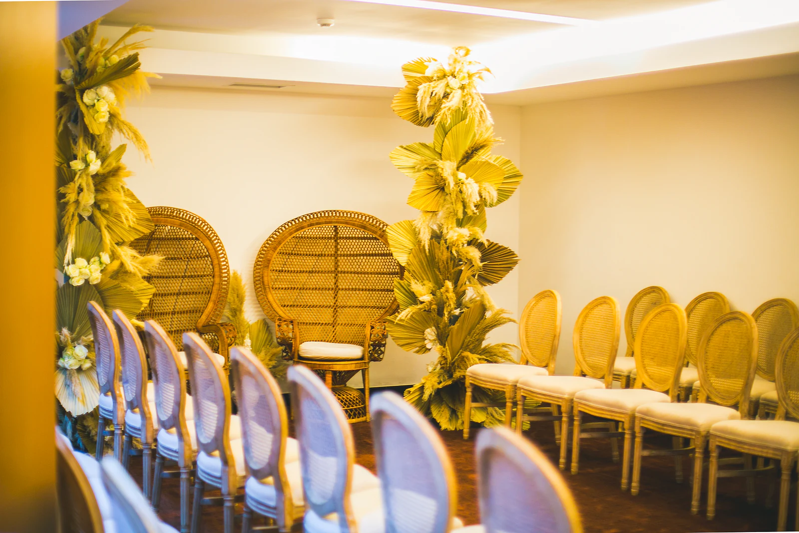 Fileira de cadeiras de palhinha com almofada branca diante de uma coluna de folhas secas e flores e das cadeiras de vime, em luz dourada