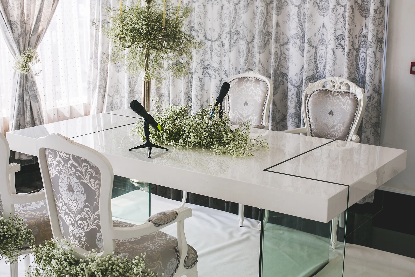 Mesa branca de tampo brilhante com microfones e gipsófila, e duas cadeiras estofadas a cinzento e branco diante de um cortinado estampado