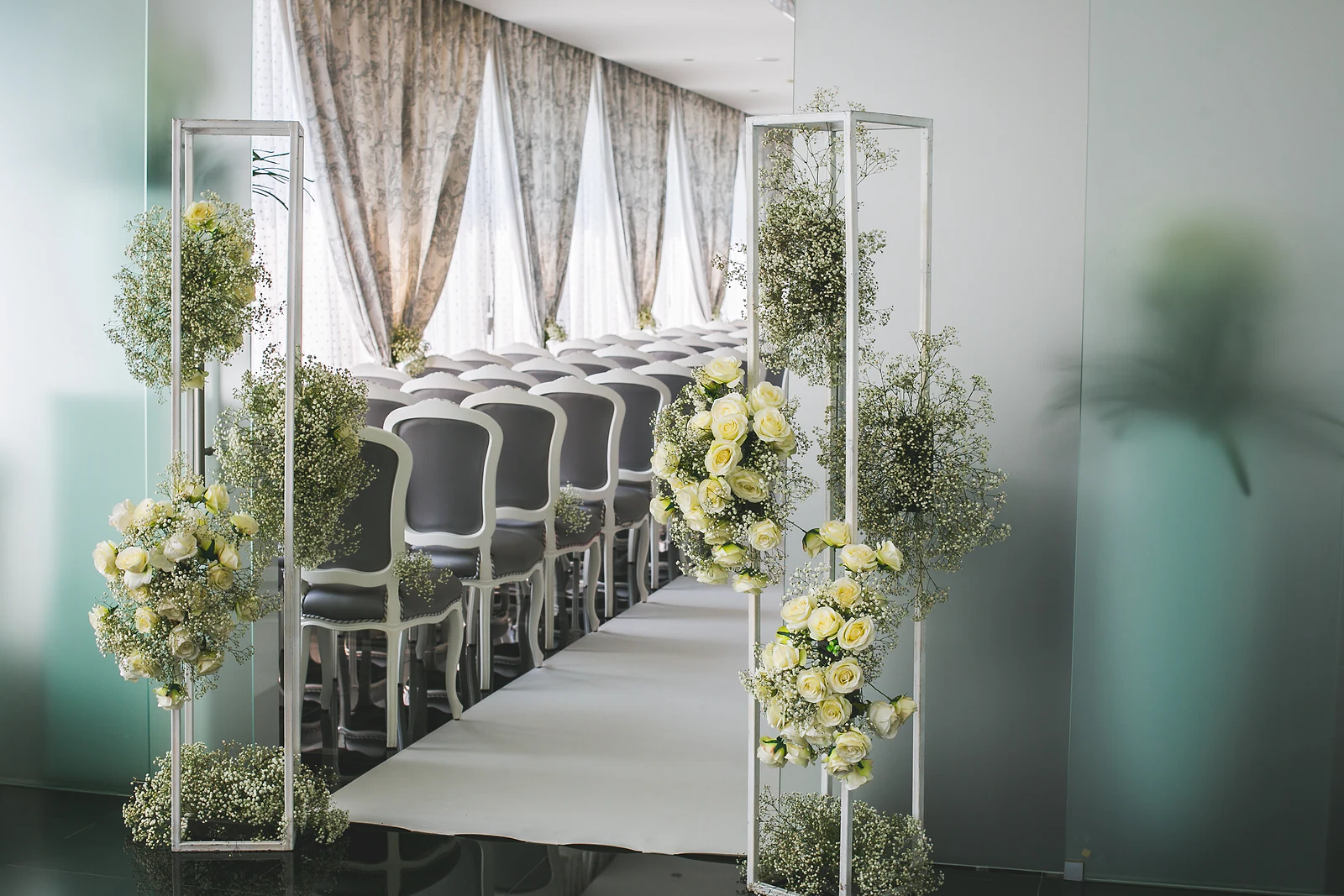 Entrada da nave ladeada por estruturas brancas com rosas cor de creme e gipsófila, diante de uma parede de vidro fosco