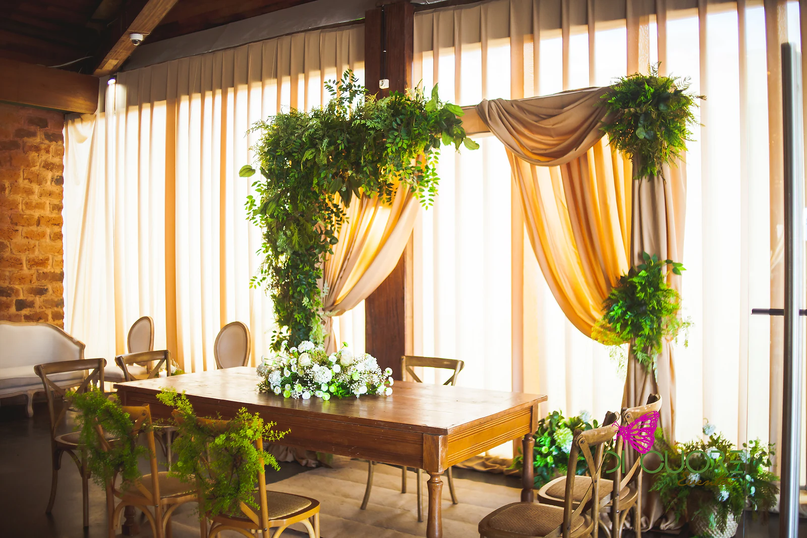 Mesa de madeira com um ramo de flores brancas diante de um arco de cortinados cor de areia e folhagem, com cortinas claras e uma parede de tijolo