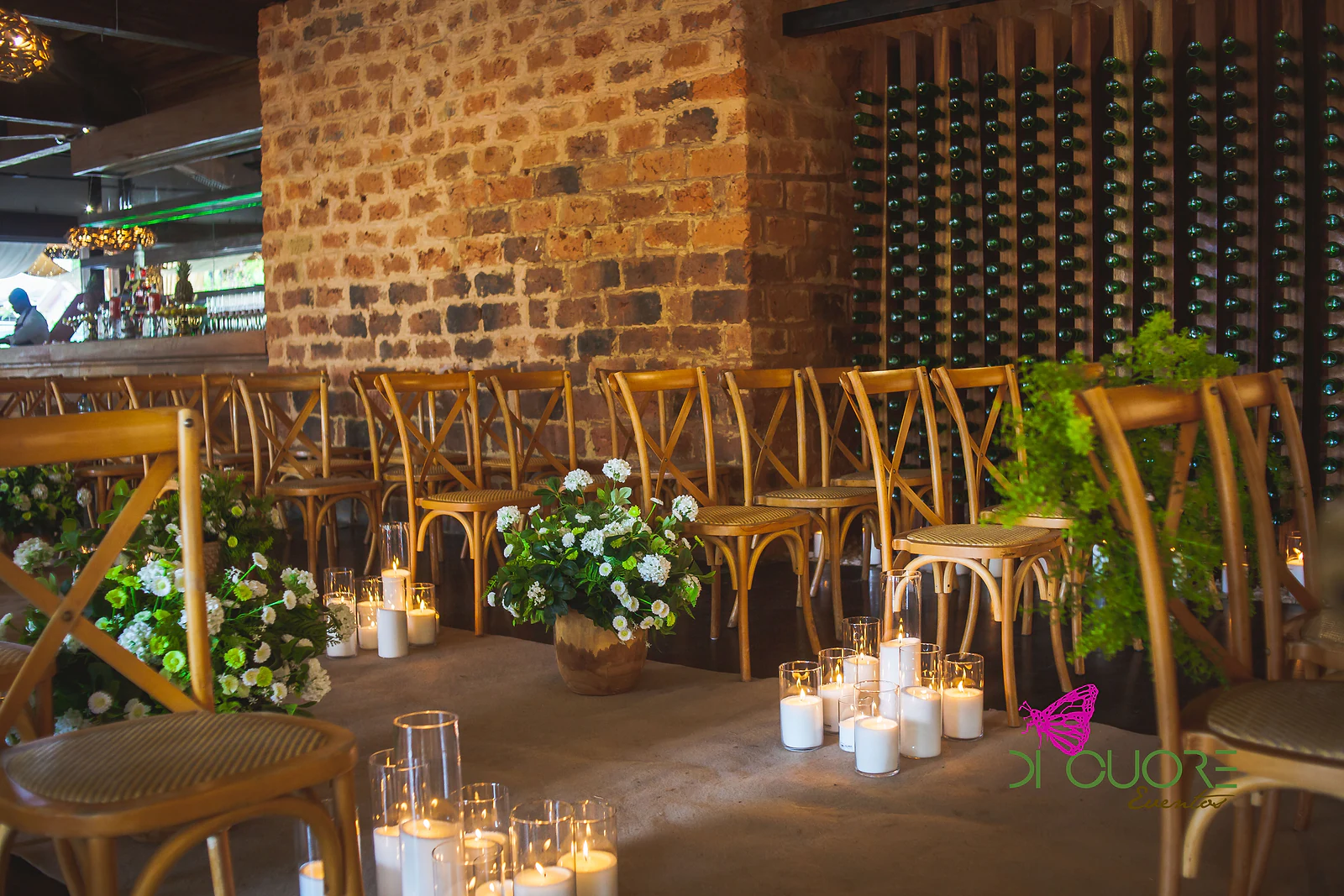 Vaso de flores brancas e folhagem com velas em cilindros de vidro ao longo da passadeira, entre cadeiras de madeira e uma parede de tijolo