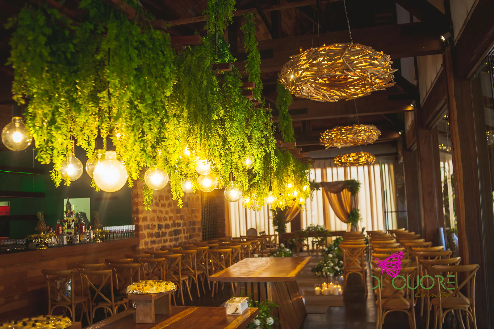 Trepadeiras e globos de luz pendurados sobre a sala, com candeeiros de ramos entrelaçados, uma mesa de madeira e cadeiras alinhadas