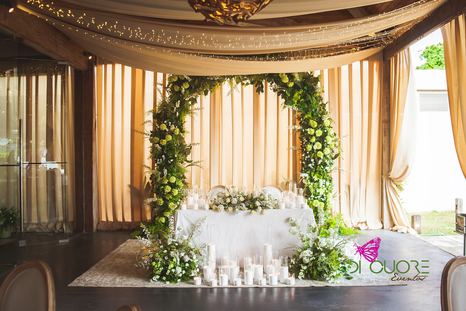 Arco de folhagem e flores brancas e verdes diante de cortinados cor de areia, com uma mesa de toalha branca, velas e flores sobre um tapete
