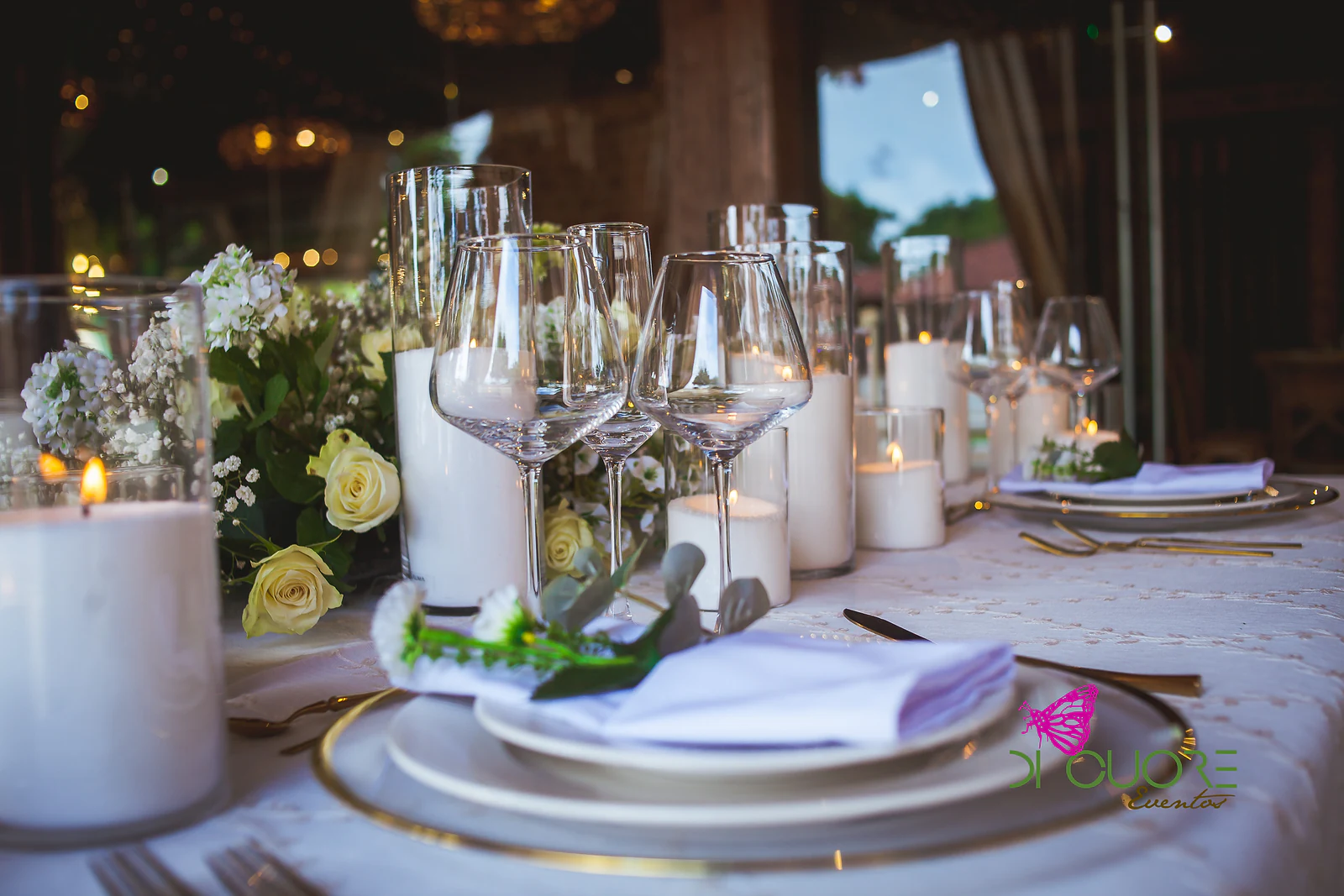 Mesa posta com copos de cristal, velas em cilindros de vidro, rosas cor de creme e guardanapo lilás sobre prato de rebordo dourado