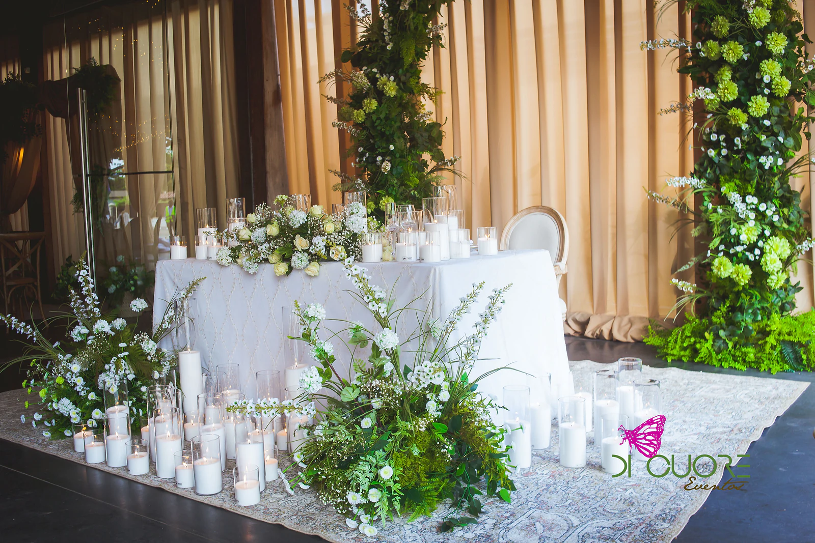 Mesa de toalha branca bordada com um ramo de rosas, rodeada de velas em cilindros de vidro e arranjos de flores brancas e folhagem