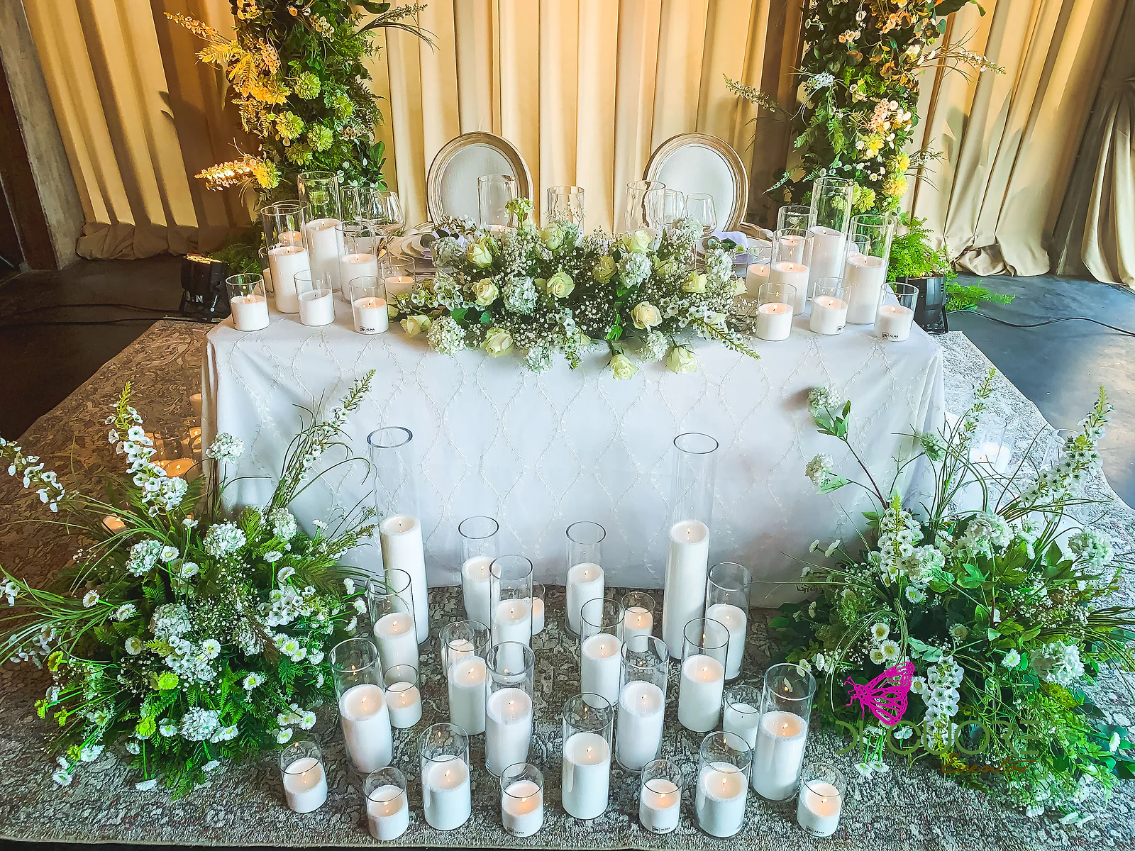 Mesa com toalha branca, velas de várias alturas e arranjos de flores brancas e folhagem, em frente a cortinados cor de creme