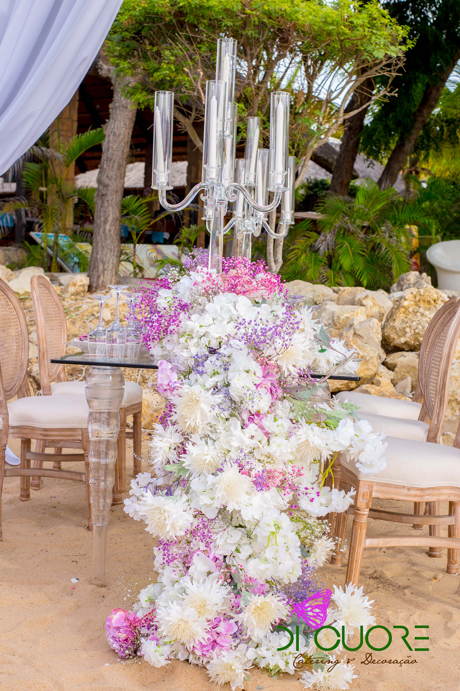 Caminho de flores brancas, rosa e lilás sobre uma mesa de pernas transparentes, com um candelabro de vidro e folhagem ao fundo