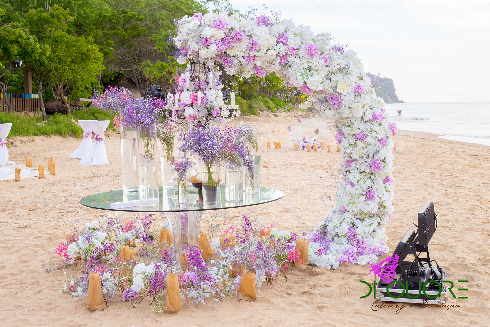 Arco de flores brancas e lilás junto a uma mesa redonda de vidro com jarras e um candelabro, sobre a areia da praia