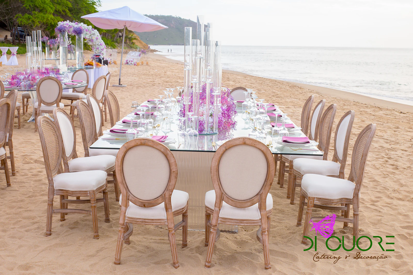 Mesa corrida de vidro posta, com cadeiras de madeira e almofadas brancas, ao longo da praia, com candelabros e flores lilás ao centro