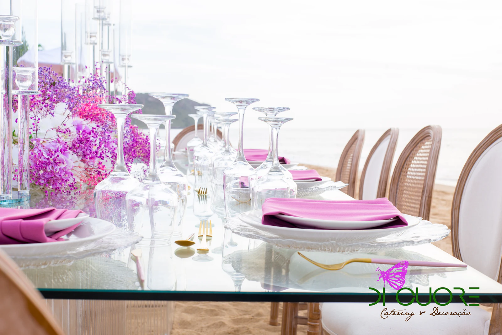 Mesa de vidro posta com guardanapos rosa, copos e talheres dourados, ao longo de um caminho de flores lilás e rosa, com o mar ao fundo