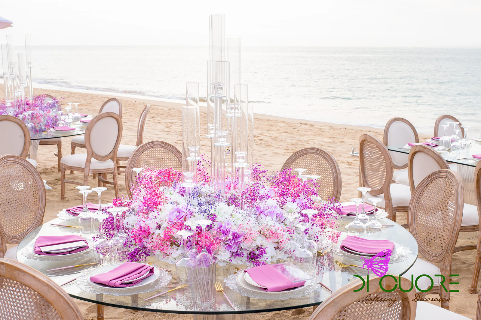 Mesa redonda de vidro com centro de flores rosa, lilás e brancas e candelabros transparentes, rodeada de cadeiras de espaldar redondo junto ao mar