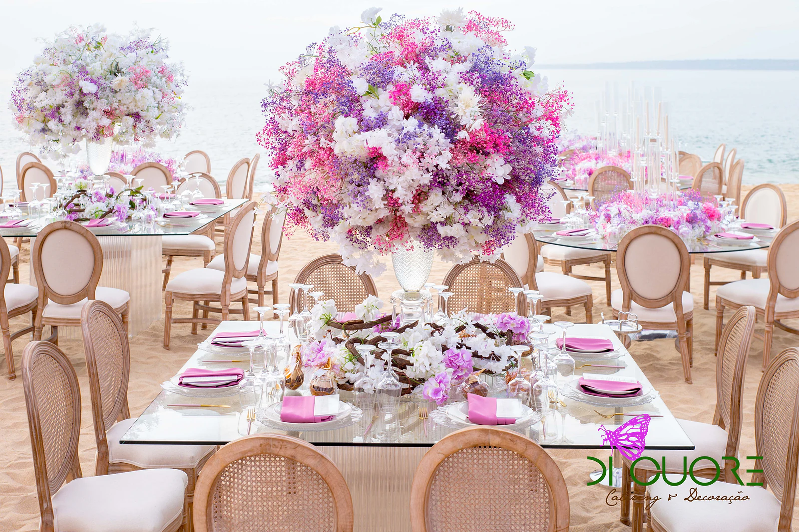 Mesa de vidro na areia da praia, com cadeiras de palhinha e um grande arranjo de flores em rosa, lilás e branco, e o mar ao fundo