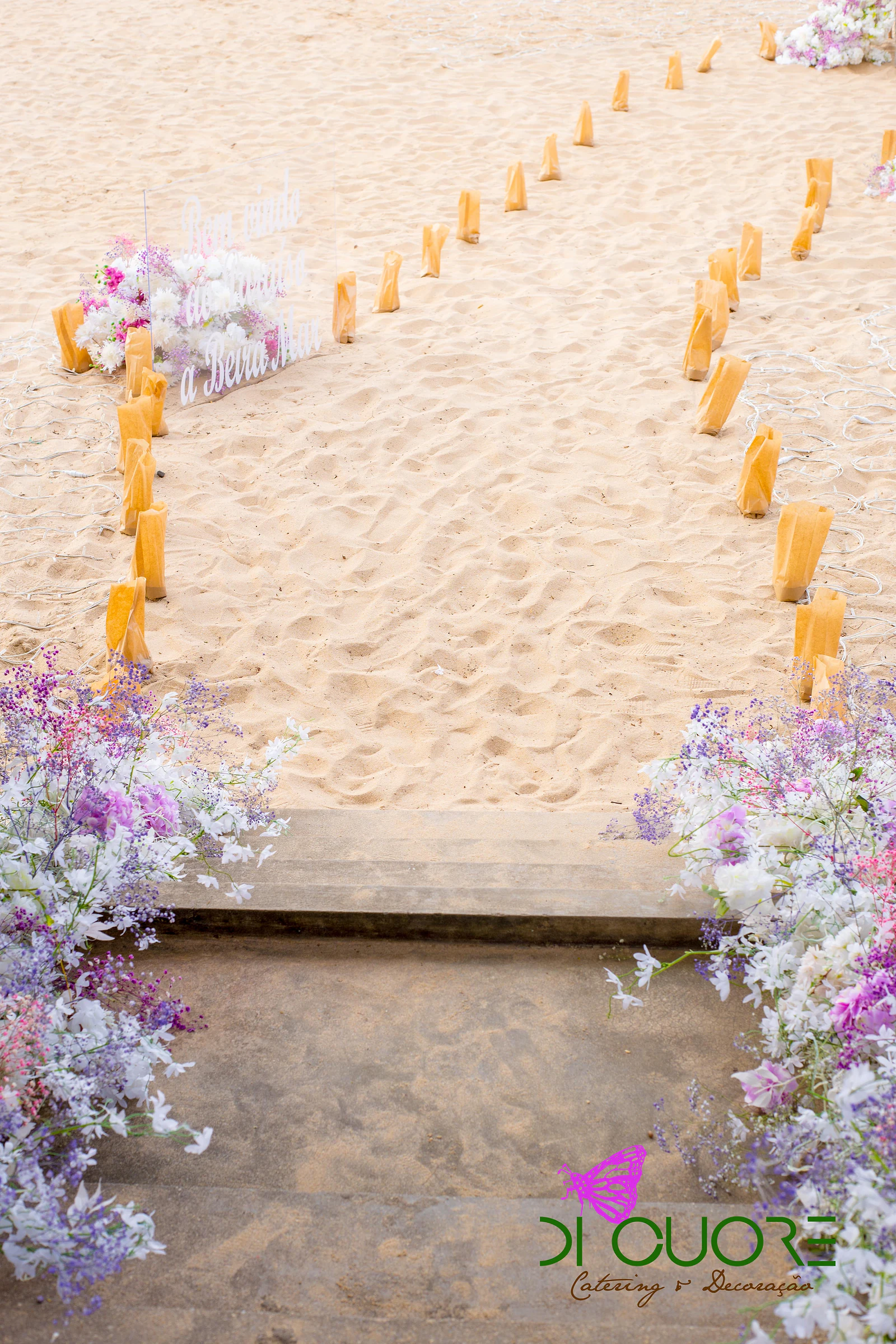 Caminho de areia marcado por saquinhos de papel dourado, entre maciços de flores lilás e brancas junto a um degrau