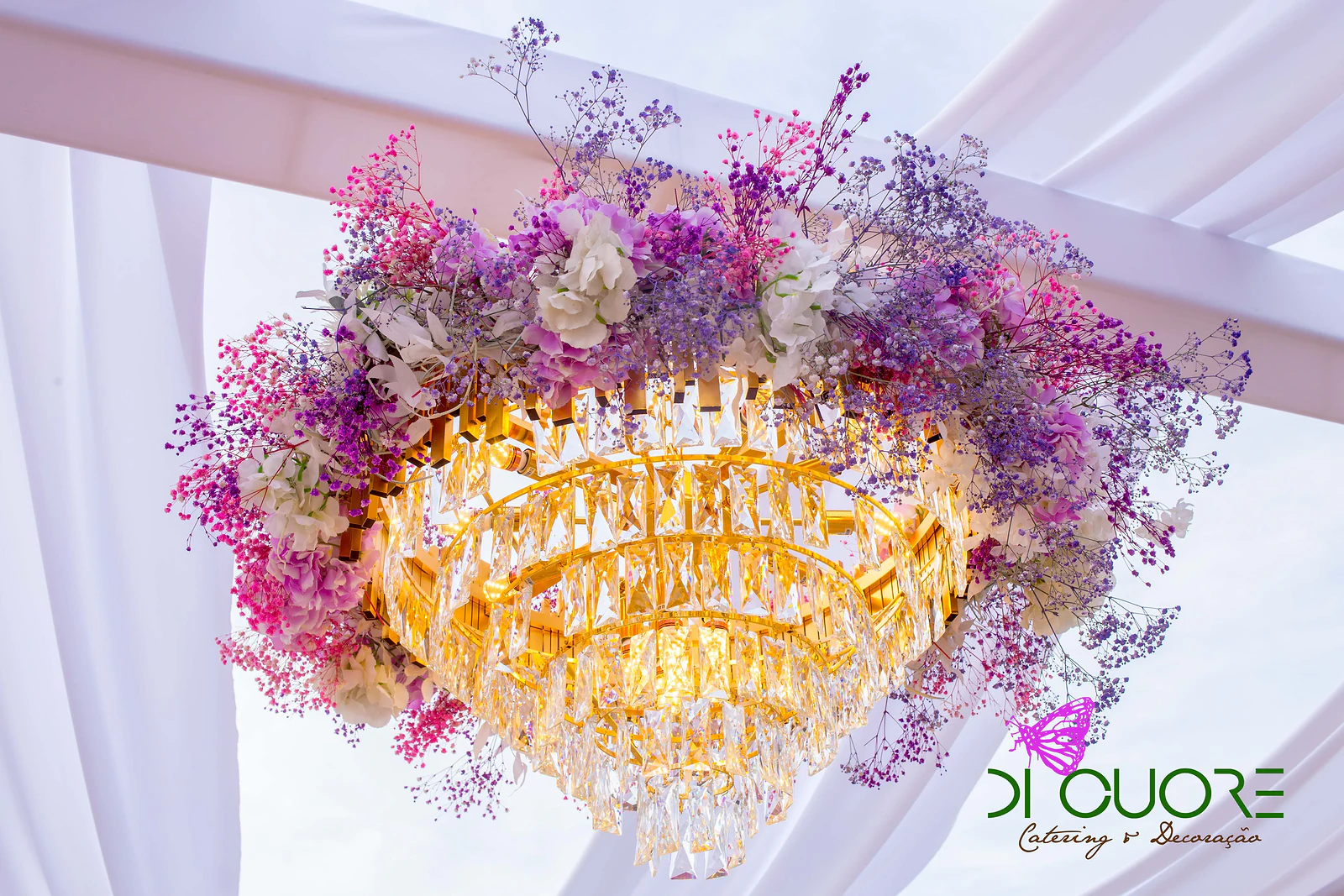 Lustre dourado de cristal envolto em gipsófila e flores rosa e lilás, suspenso entre cortinados brancos