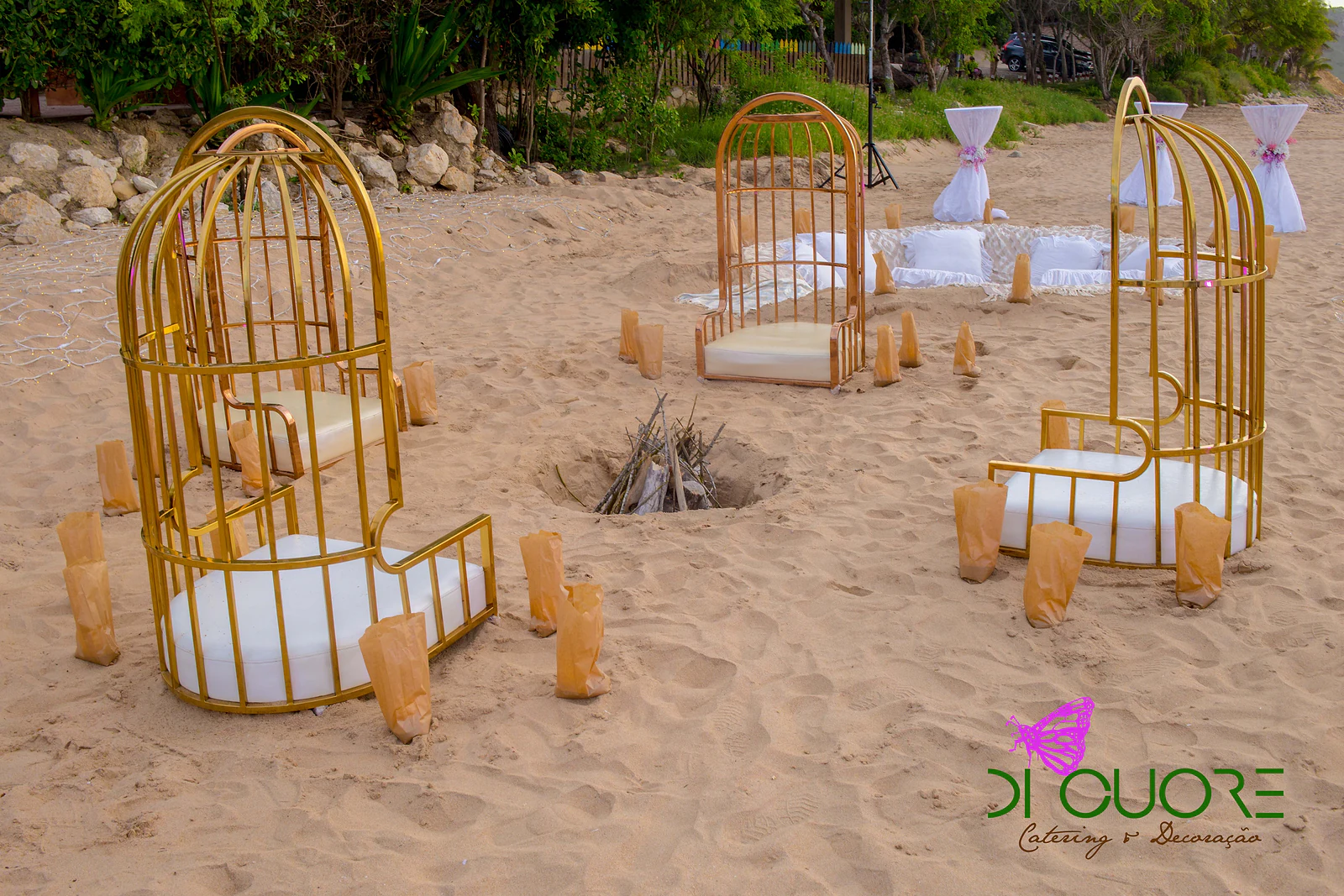 Gaiolas douradas com almofadas brancas e saquinhos de papel dourado dispostos em círculo na areia, com uma fogueira montada ao centro