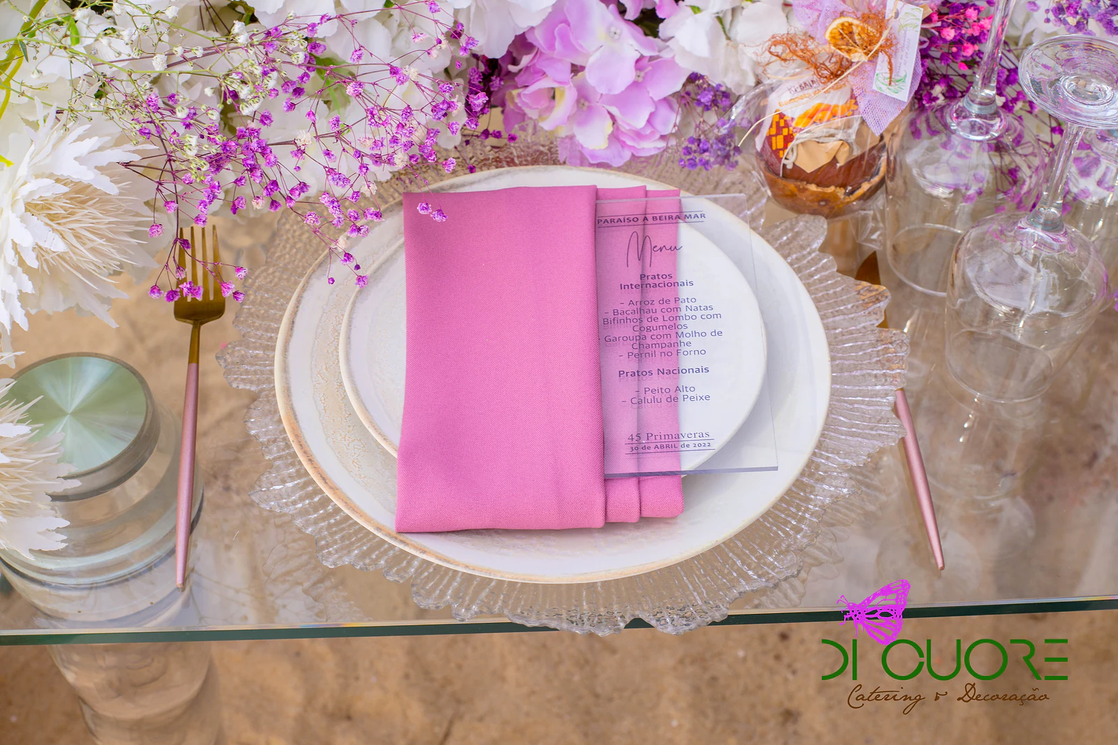 Lugar posto sobre tampo de vidro, com prato de vidro trabalhado, guardanapo rosa, menu e talheres dourados entre flores lilás