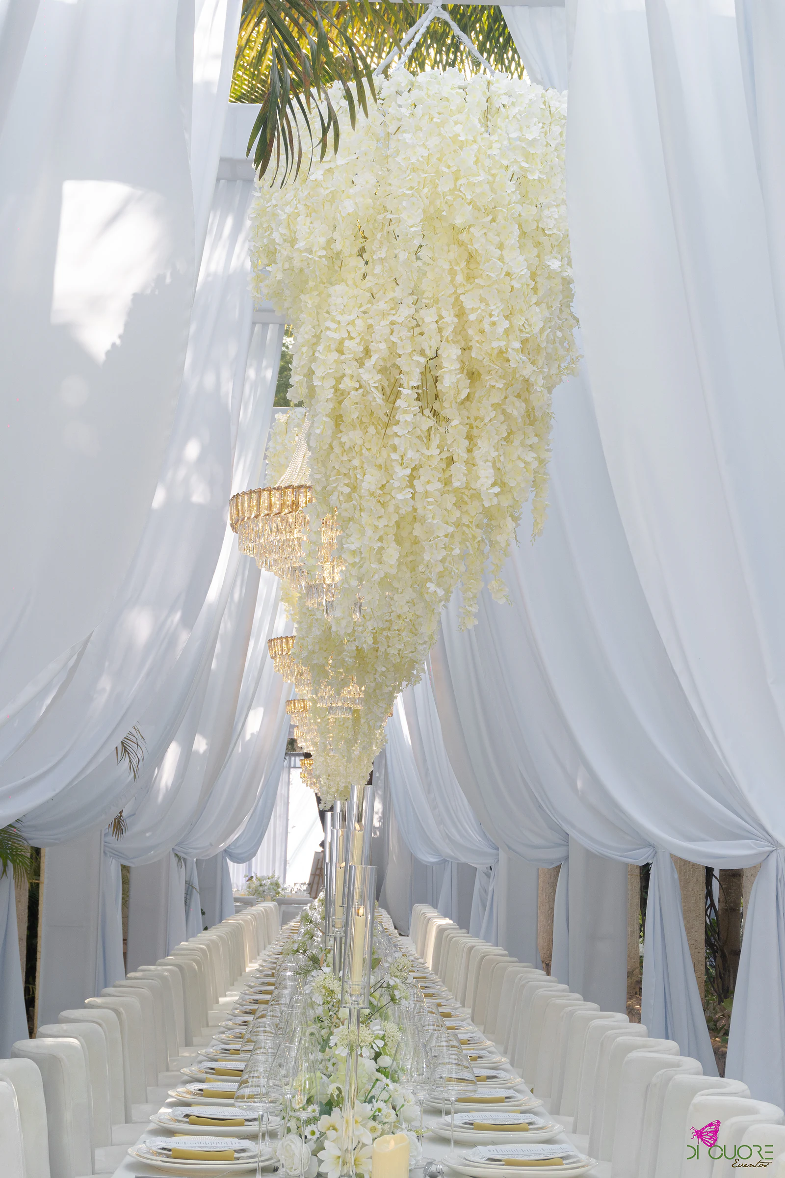 Cascata de flores brancas suspensa sobre a mesa corrida, entre cortinados soltos, com lustres de cristal por baixo