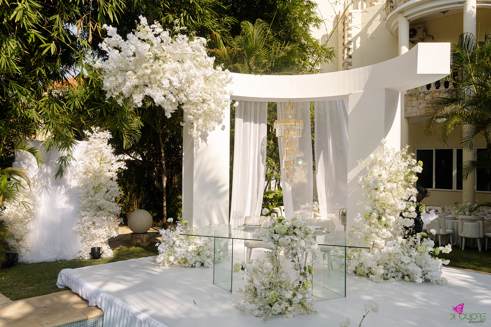 Plataforma branca ao ar livre com pórtico curvo, cortinas leves e lustre dourado, mesa de vidro e maciços de flores brancas dos dois lados