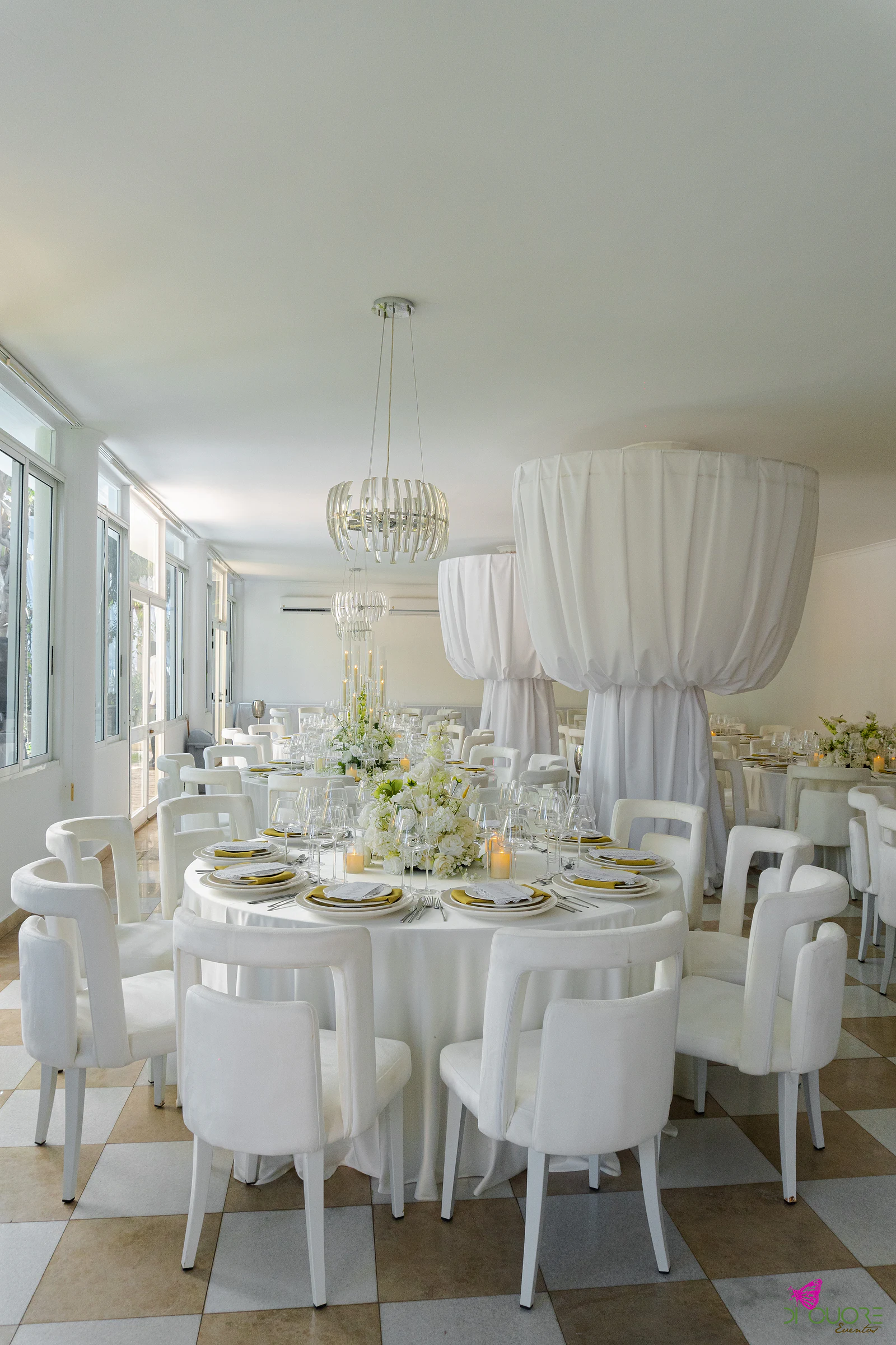 Salão com mesa redonda posta a branco e dourado, flores brancas, cadeiras brancas, lustres e colunas de tecido sobre um chão axadrezado