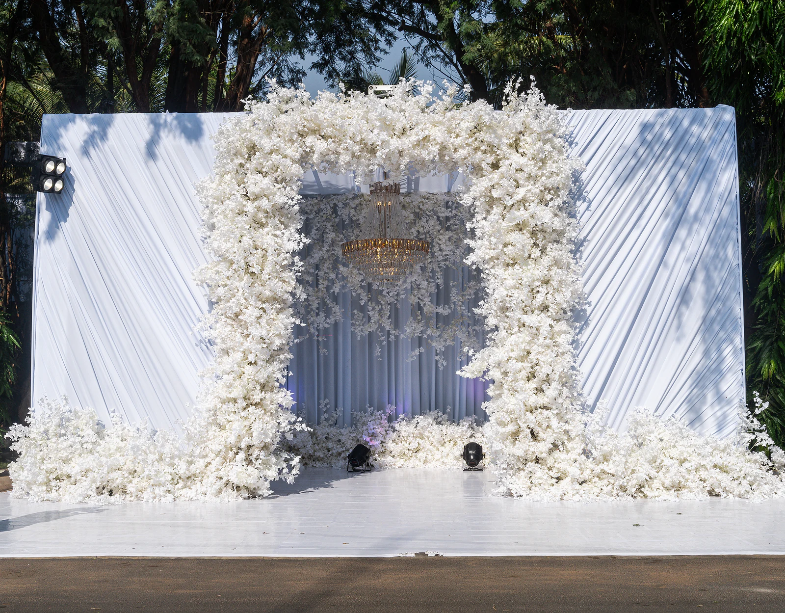 Painel de tecido branco plissado com um arco de flores brancas e um lustre dourado ao centro
