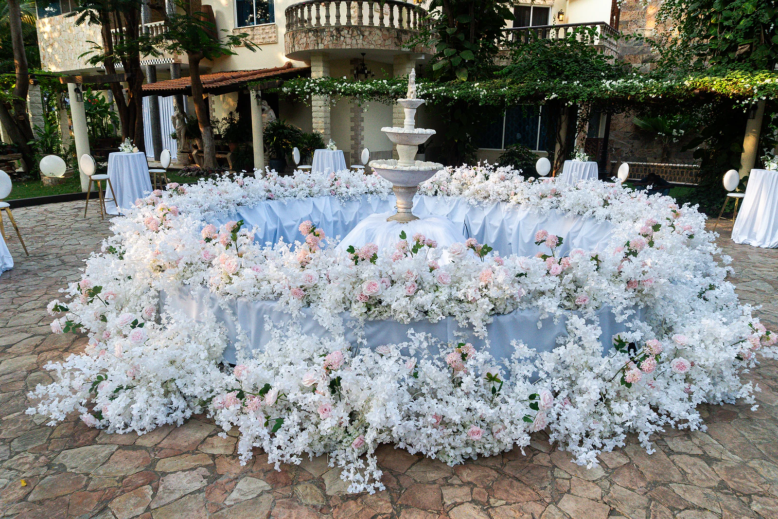 Fonte de jardim rodeada por uma coroa de flores brancas e cor-de-rosa pálido sobre tecido azul-acinzentado, num pátio de pedra
