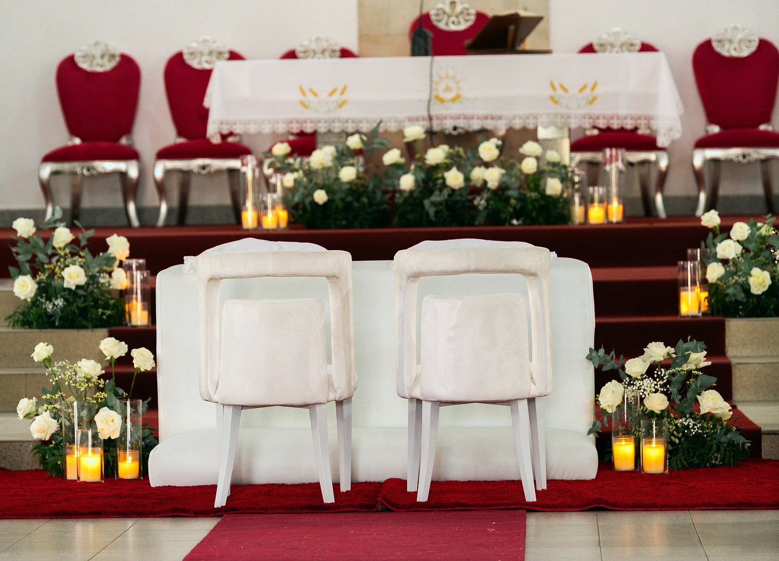 Duas cadeiras brancas diante do altar, com degraus de tapete vermelho, rosas brancas e velas em cilindros de vidro e cadeiras vermelhas ao fundo