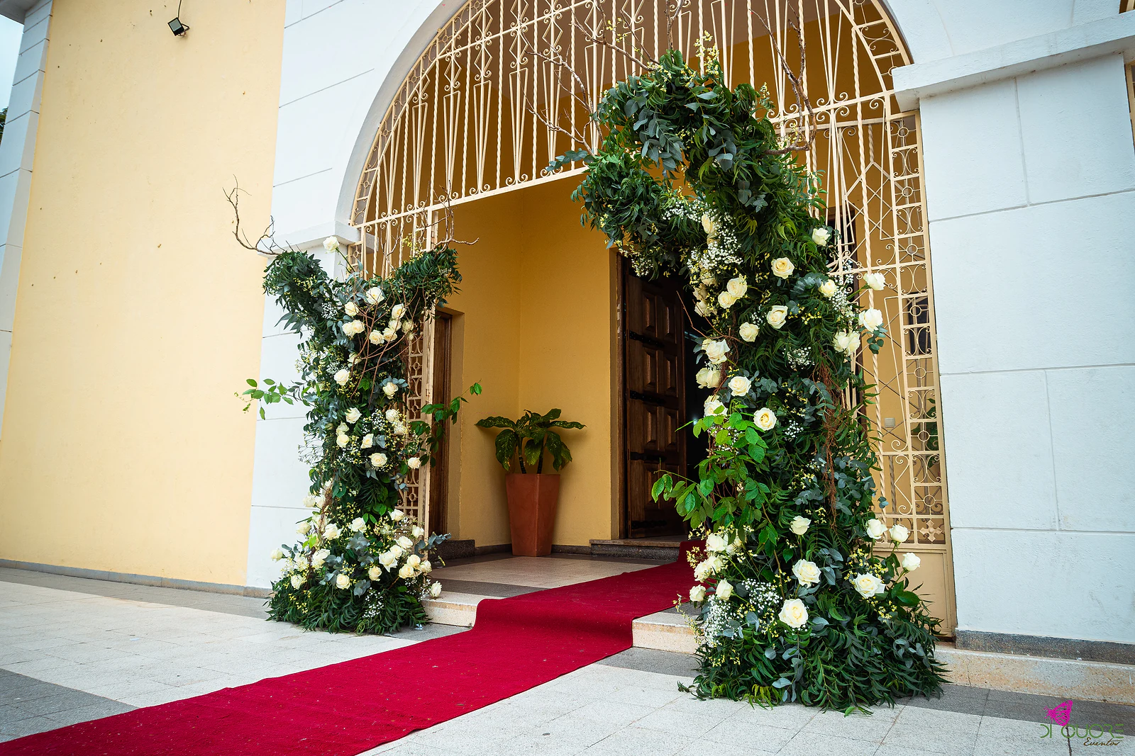 Portal da igreja visto de lado, com uma coluna de rosas brancas e folhagem a subir pela grade dourada e o tapete vermelho em diagonal