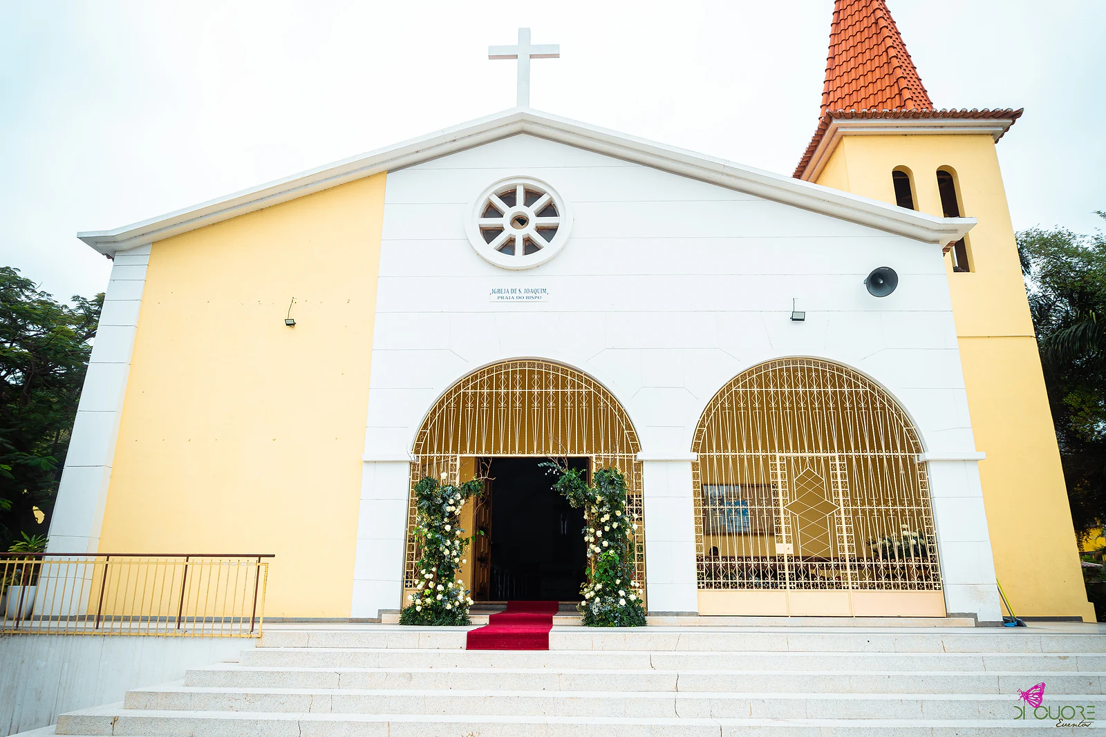 Fachada da igreja em amarelo e branco, com cruz, janela redonda e campanário de telhado vermelho; no topo da escadaria, o portal com colunas de flores e tapete vermelho