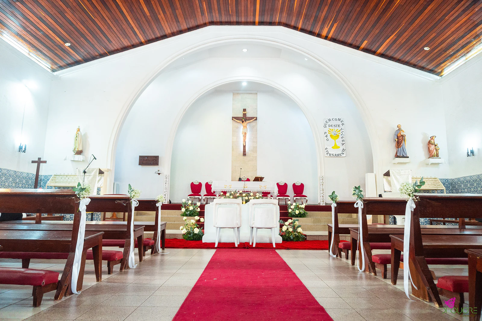 Nave da igreja com corredor de tapete vermelho, laços de fita branca nos bancos e as cadeiras brancas dos noivos ao fundo