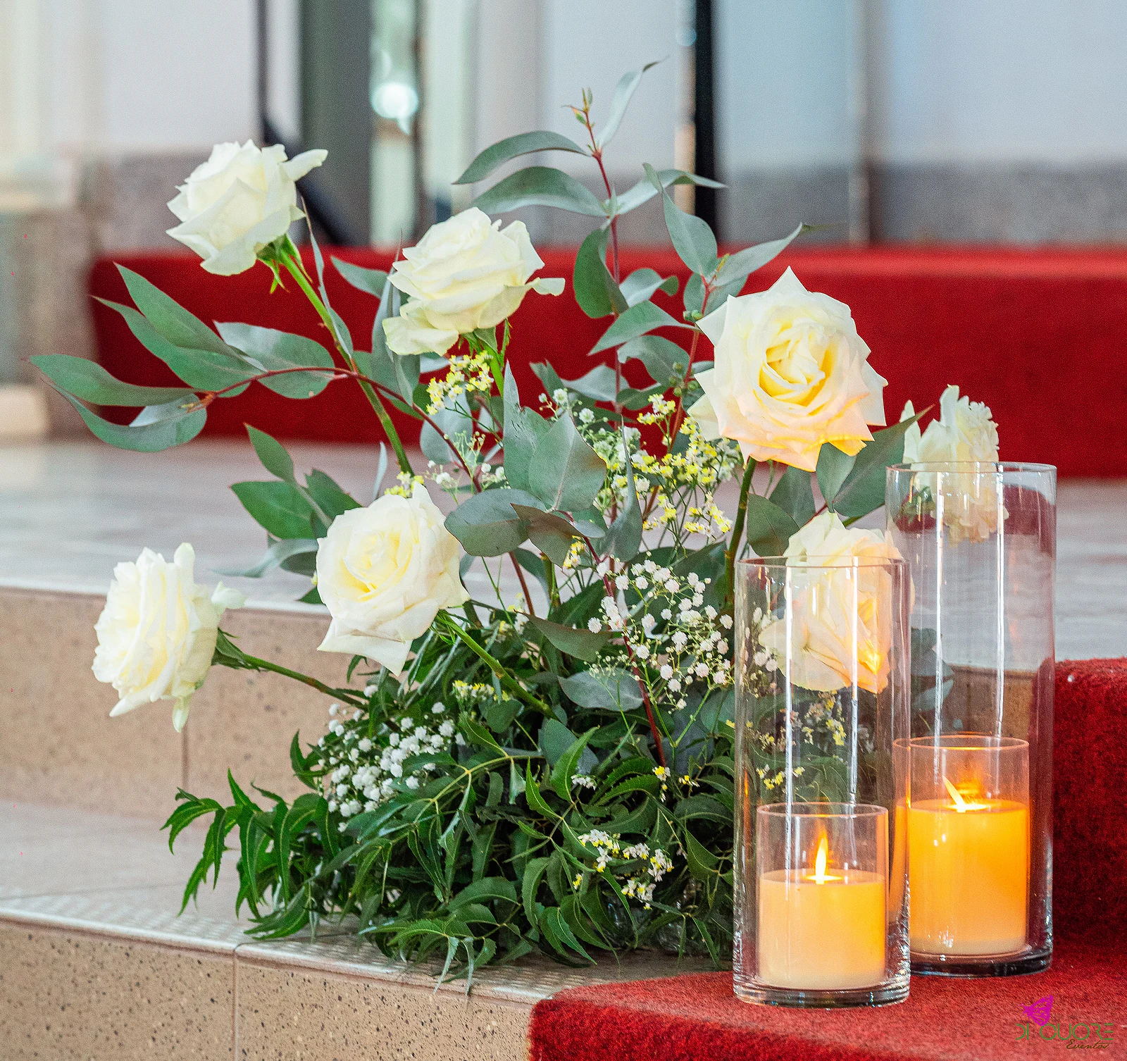 Rosas brancas, gipsófila e folhagem junto a cilindros de vidro com velas acesas, sobre um degrau de pedra e tapete vermelho