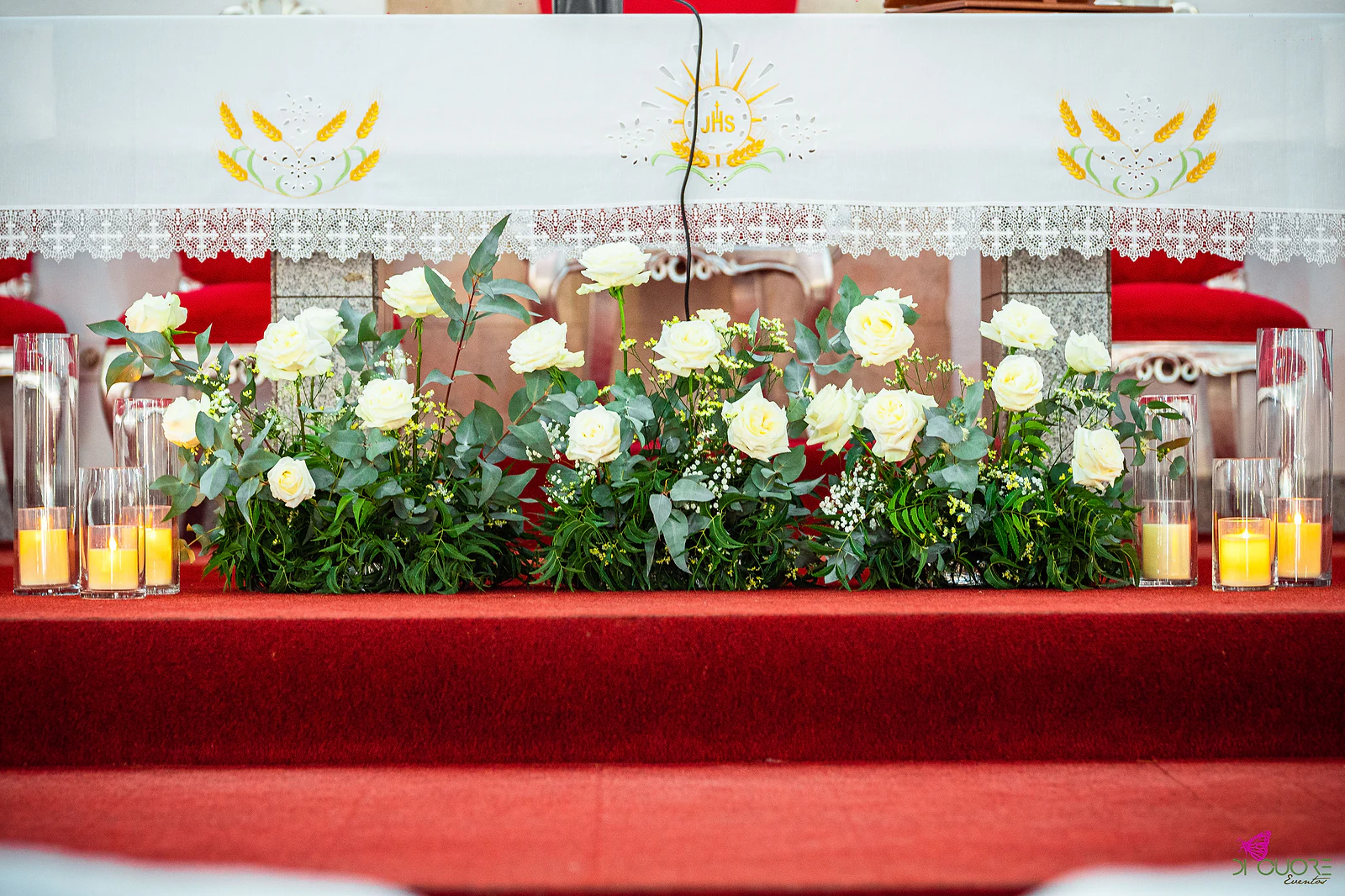 Altar com toalha rendada, rosas brancas e eucalipto e velas em cilindros de vidro sobre os degraus de tapete vermelho