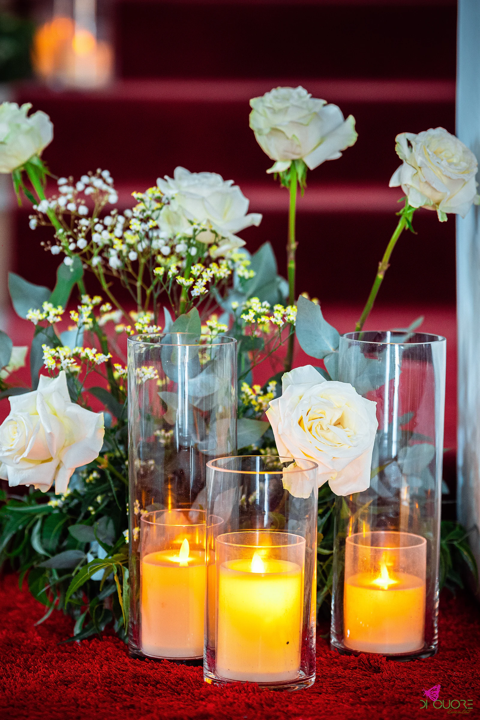 Rosas brancas, gipsófila e eucalipto com três velas em cilindros de vidro, sobre tapete vermelho