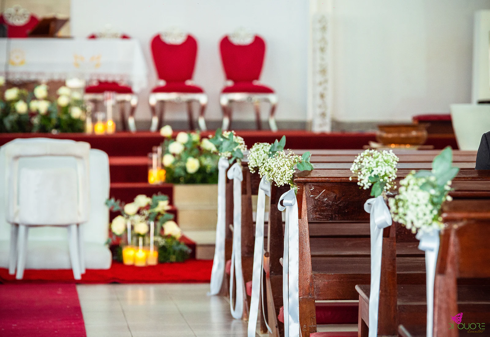 Cadeiras brancas diante do altar, com rosas brancas e velas nos degraus, e uma fila de bancos com ramos de gipsófila e laços de fita branca em primeiro plano