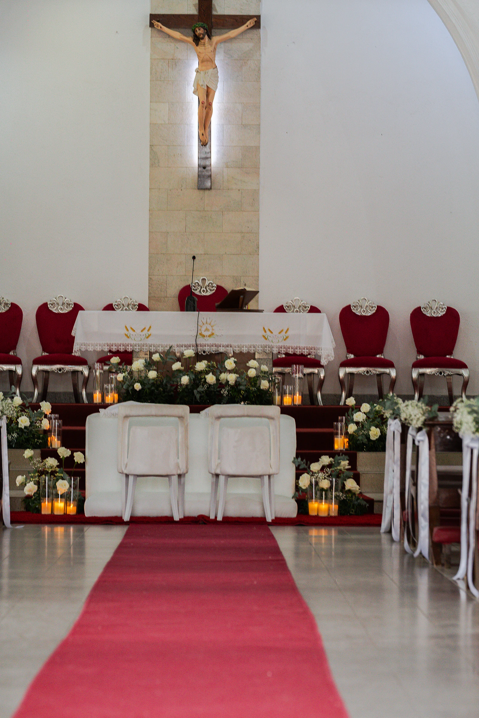 Corredor de tapete vermelho até ao altar, com as cadeiras brancas dos noivos, rosas brancas, velas e o crucifixo ao fundo