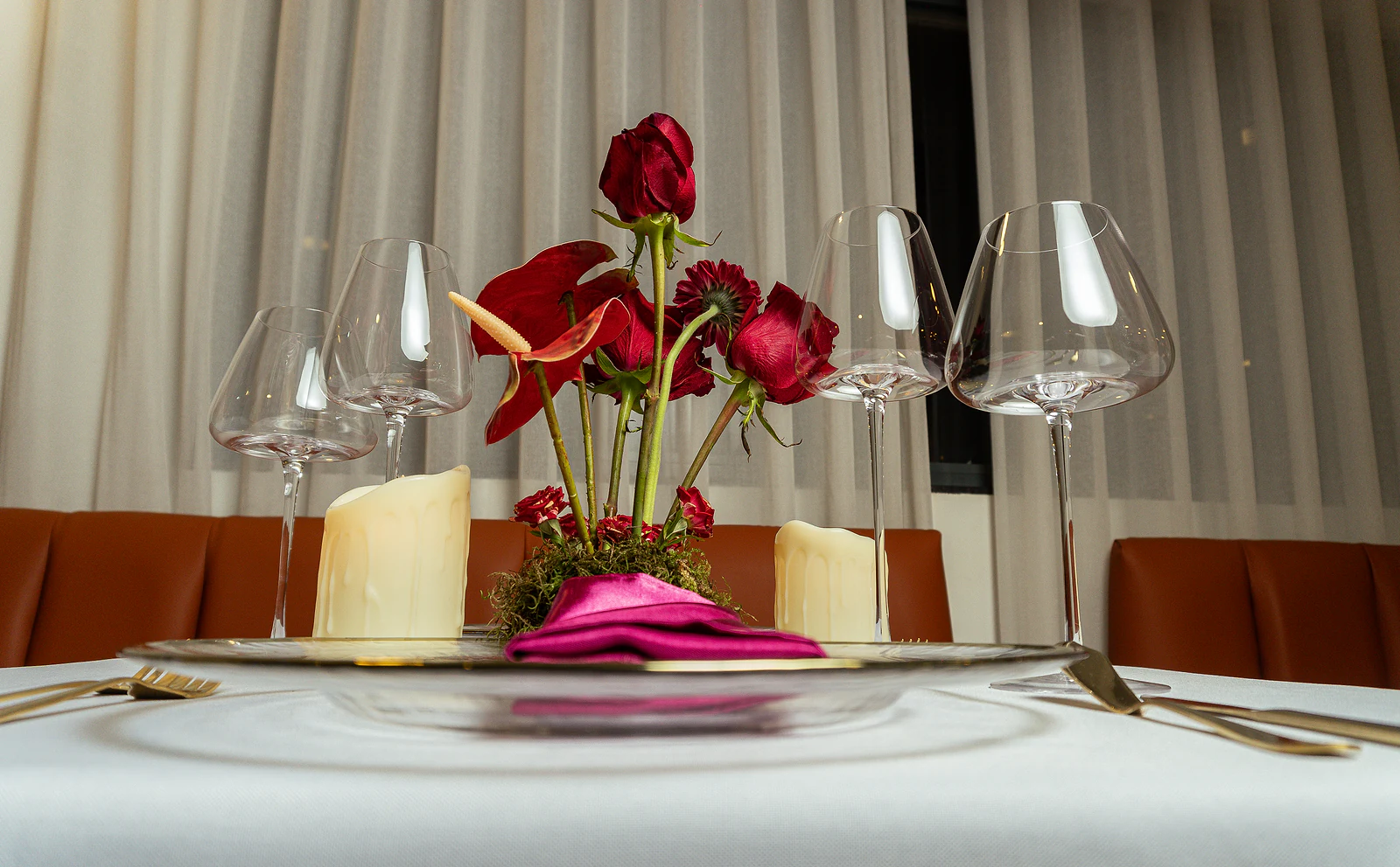 Arranjo baixo de rosas e antúrios vermelhos entre taças altas e velas, visto ao nível da mesa, com o banco cor de telha ao fundo