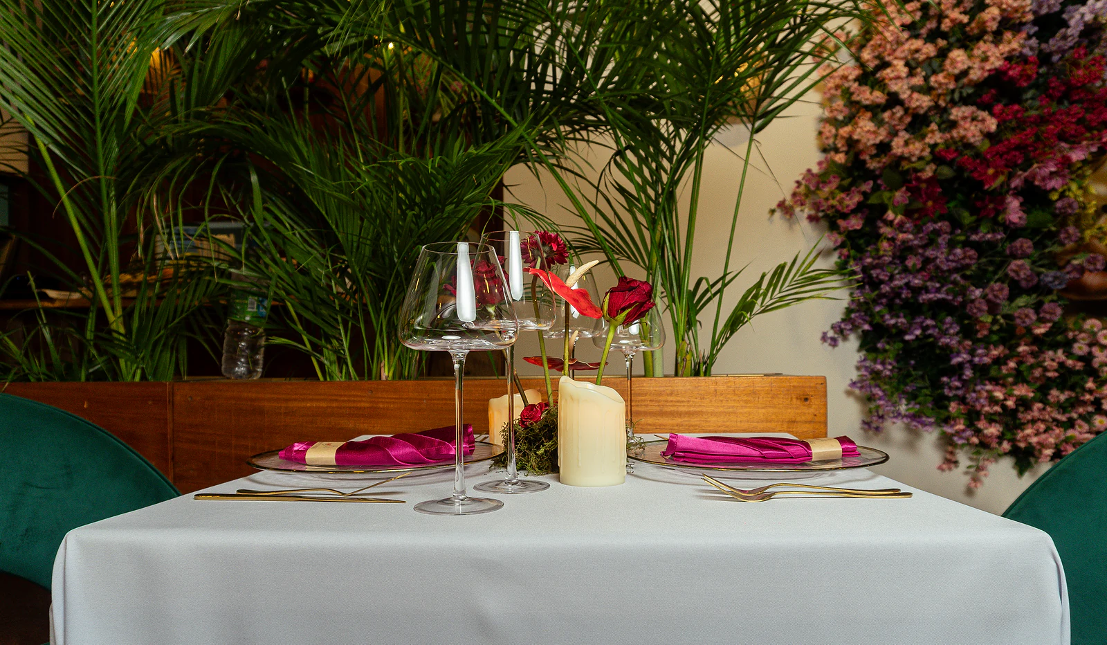 Mesa para dois diante de palmeiras e de uma parede de flores cor-de-rosa e lilases, com guardanapos fúcsia e velas