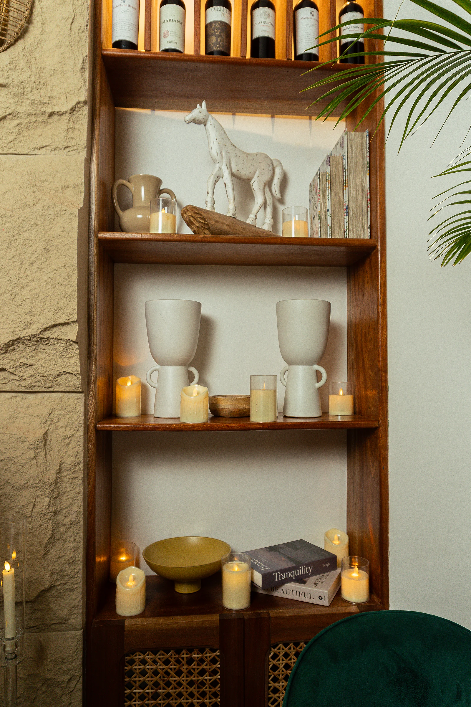 Estante de madeira com cerâmicas brancas, velas e livros, junto a uma parede de pedra