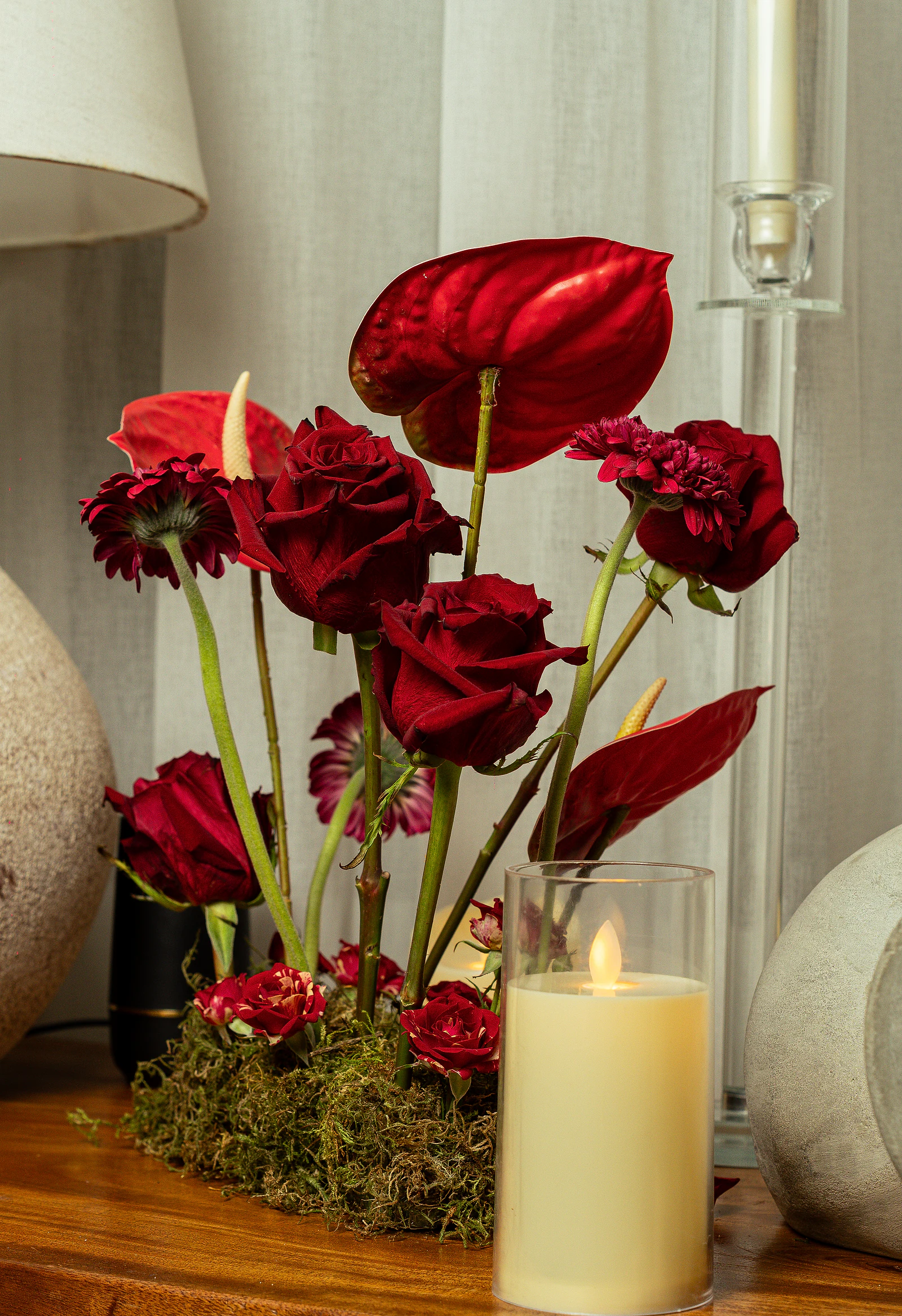 Arranjo de rosas e antúrios vermelhos sobre musgo, junto a uma vela de vidro