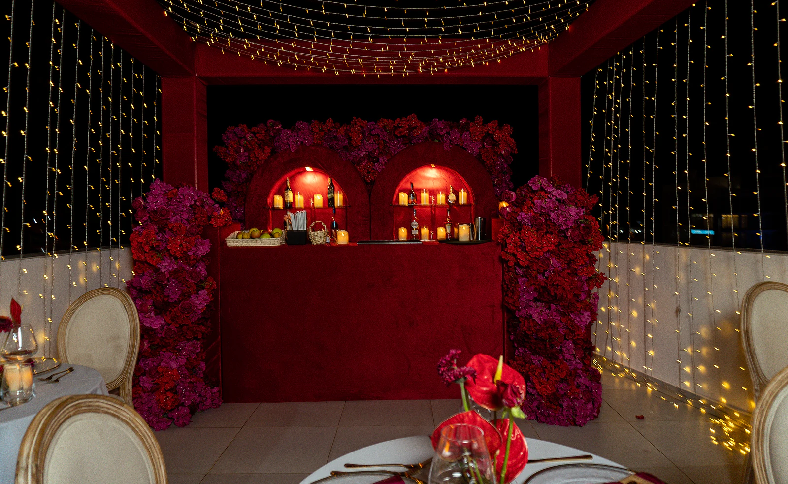 Balcão de veludo vermelho com dois nichos em arco iluminados, entre painéis de flores vermelhas e lilases, sob uma cobertura de luzes