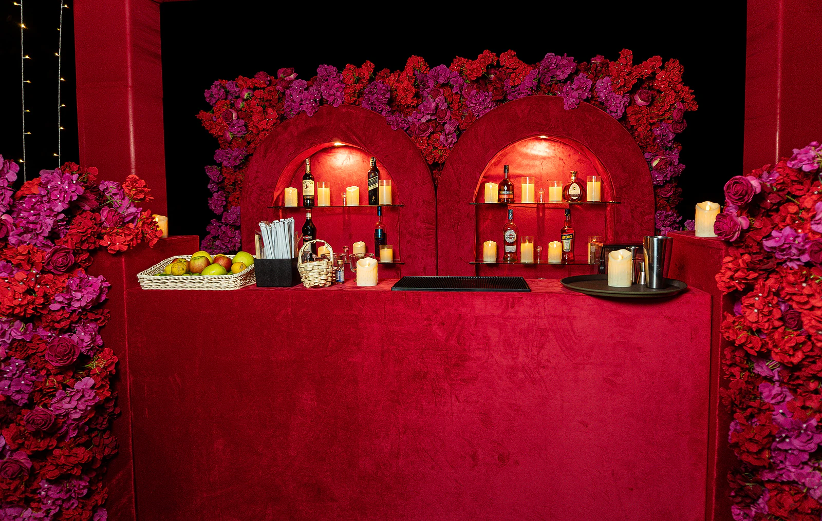 Balcão de veludo vermelho com garrafas, velas e um cesto de fruta, diante de dois nichos em arco e de flores vermelhas e lilases