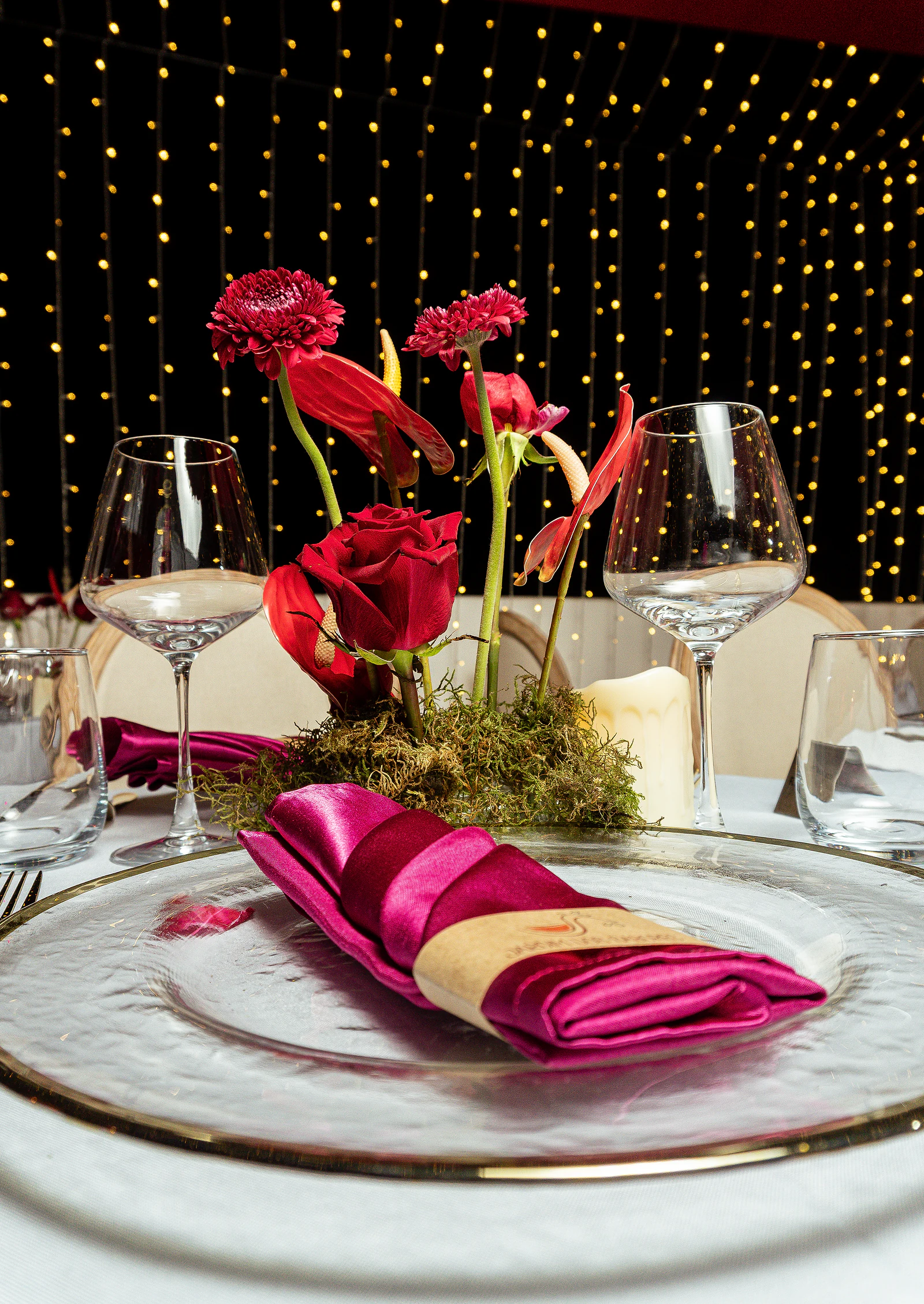 Fuchsia satin napkin on a glass plate, with an arrangement of red roses and wine glasses in front of a curtain of fairy lights