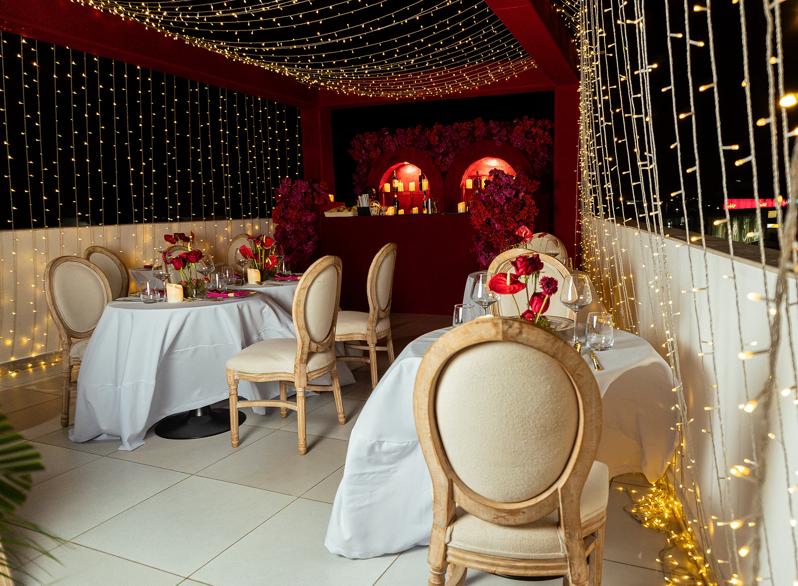 Recanto com cortinas de luzes, mesas redondas de toalha branca, cadeiras de madeira clara de costas ovais e o balcão vermelho ao fundo