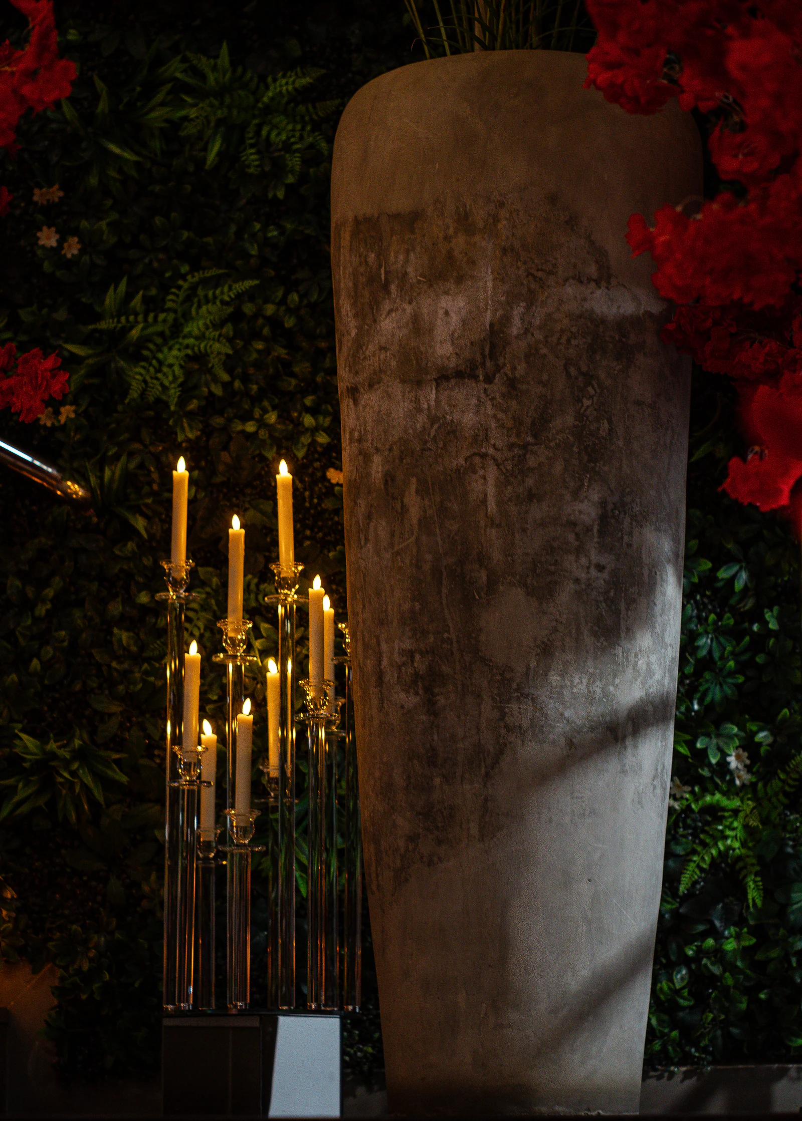 Candelabro de vidro com velas altas ao lado de um grande vaso cinzento, diante de uma parede verde, com flores vermelhas nos cantos