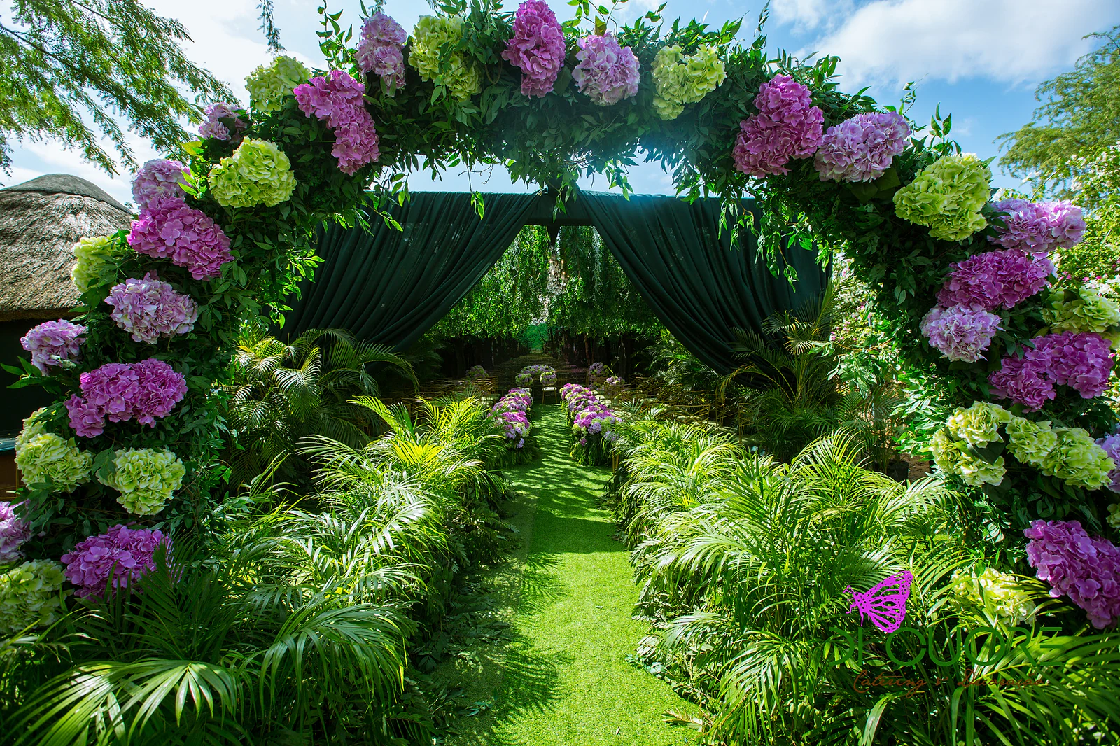 Arch of lilac, pink and lime-green hydrangeas over the grass aisle, with palms on either side and dark green curtains beyond